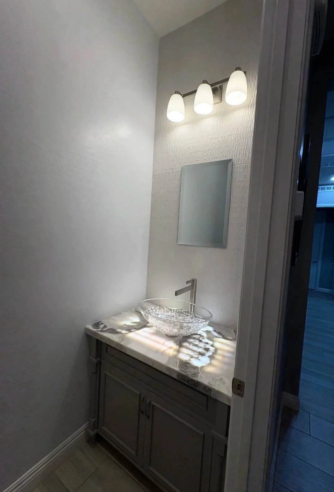 Small bathroom with a marble countertop, glass sink, mirror, and three-bulb wall light fixture.