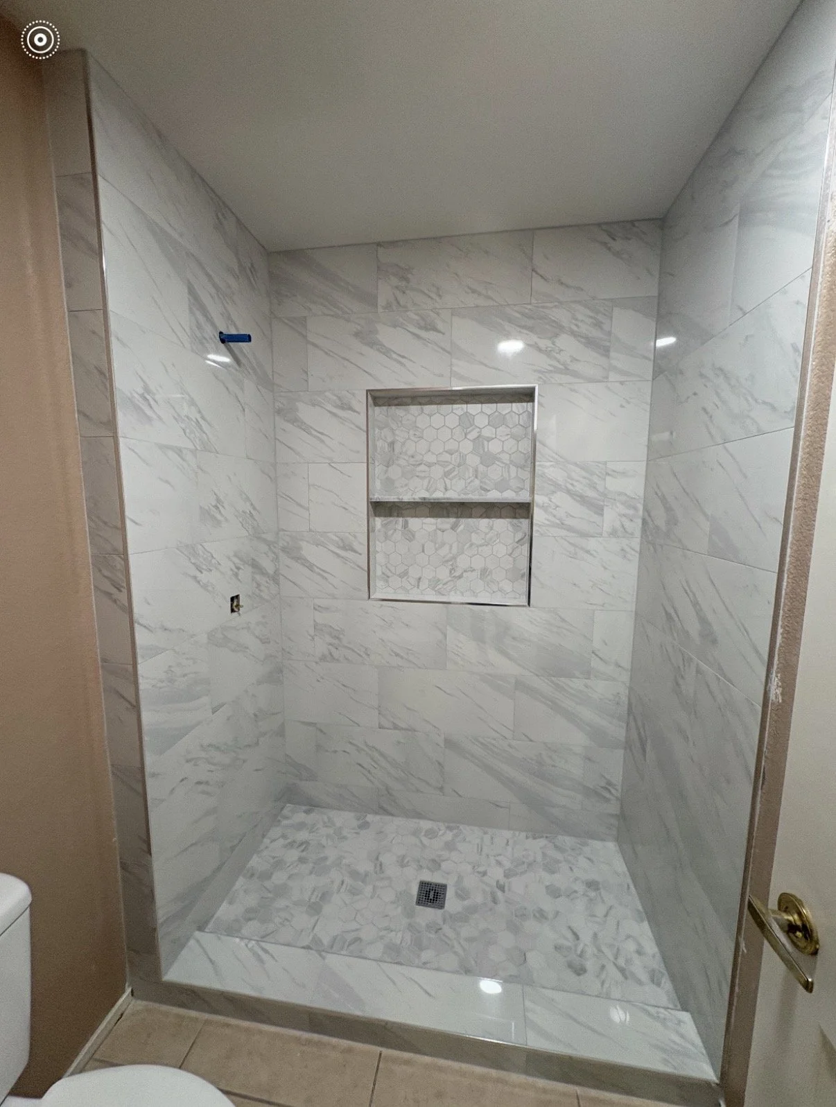 Bathroom shower with white marble tiles, a built-in niche with hexagonal patterned tiles, and a drain on the floor.