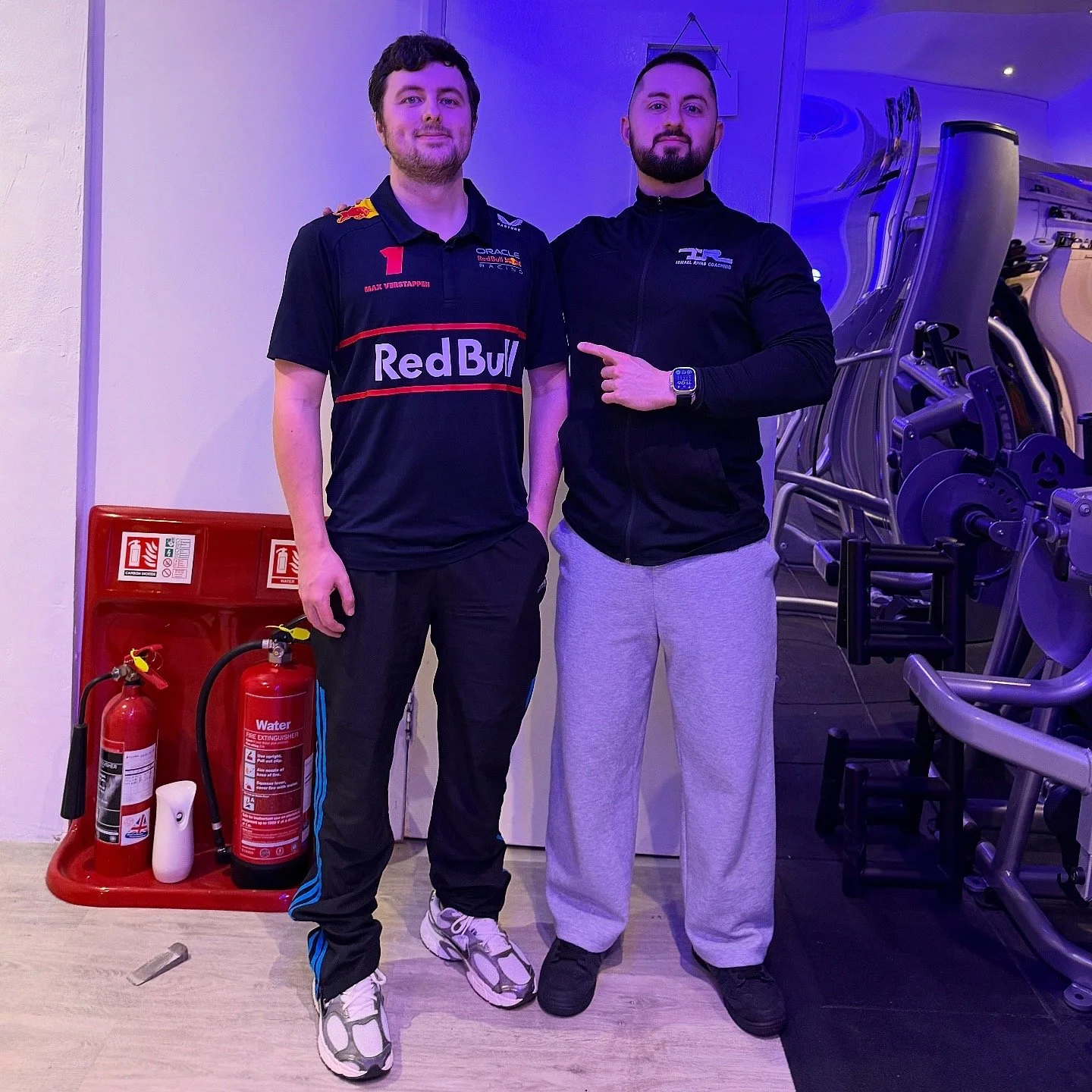 Two men standing next to each other in a fitness center. One is dressed in a Red Bull Racing team uniform, and the other is wearing a black zip-up jacket and gray sweatpants. The man in the uniform has his hand on the shoulder of the man in casual wear. There are fire extinguishers and a deletion box nearby, with gym equipment in the background.