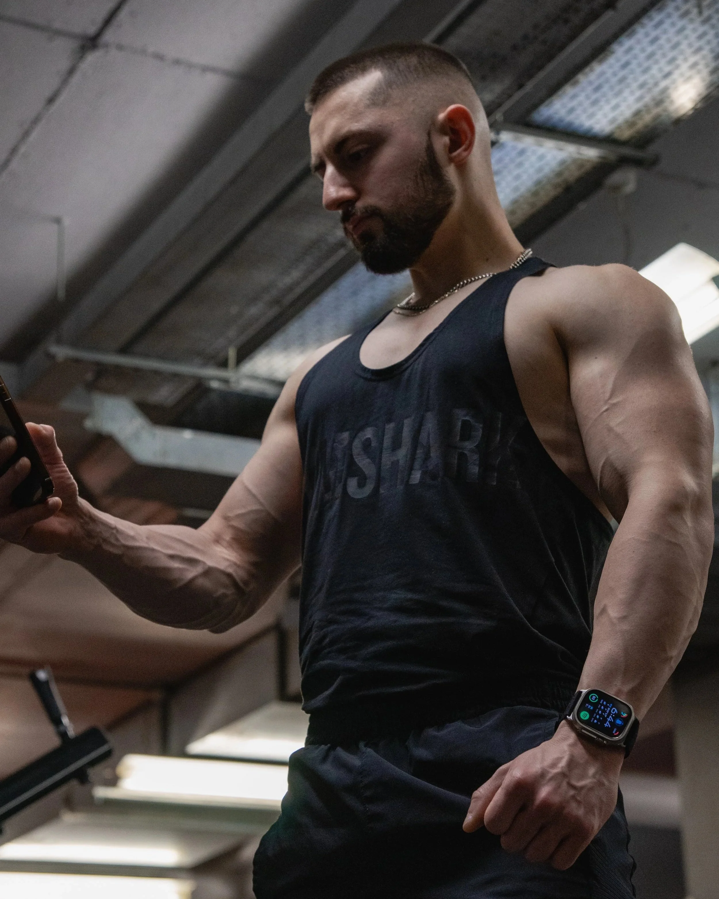A man with a beard wearing a sleeveless black shirt and a smartwatch on his left wrist in an indoor gym, looking down at his phone with a serious expression.