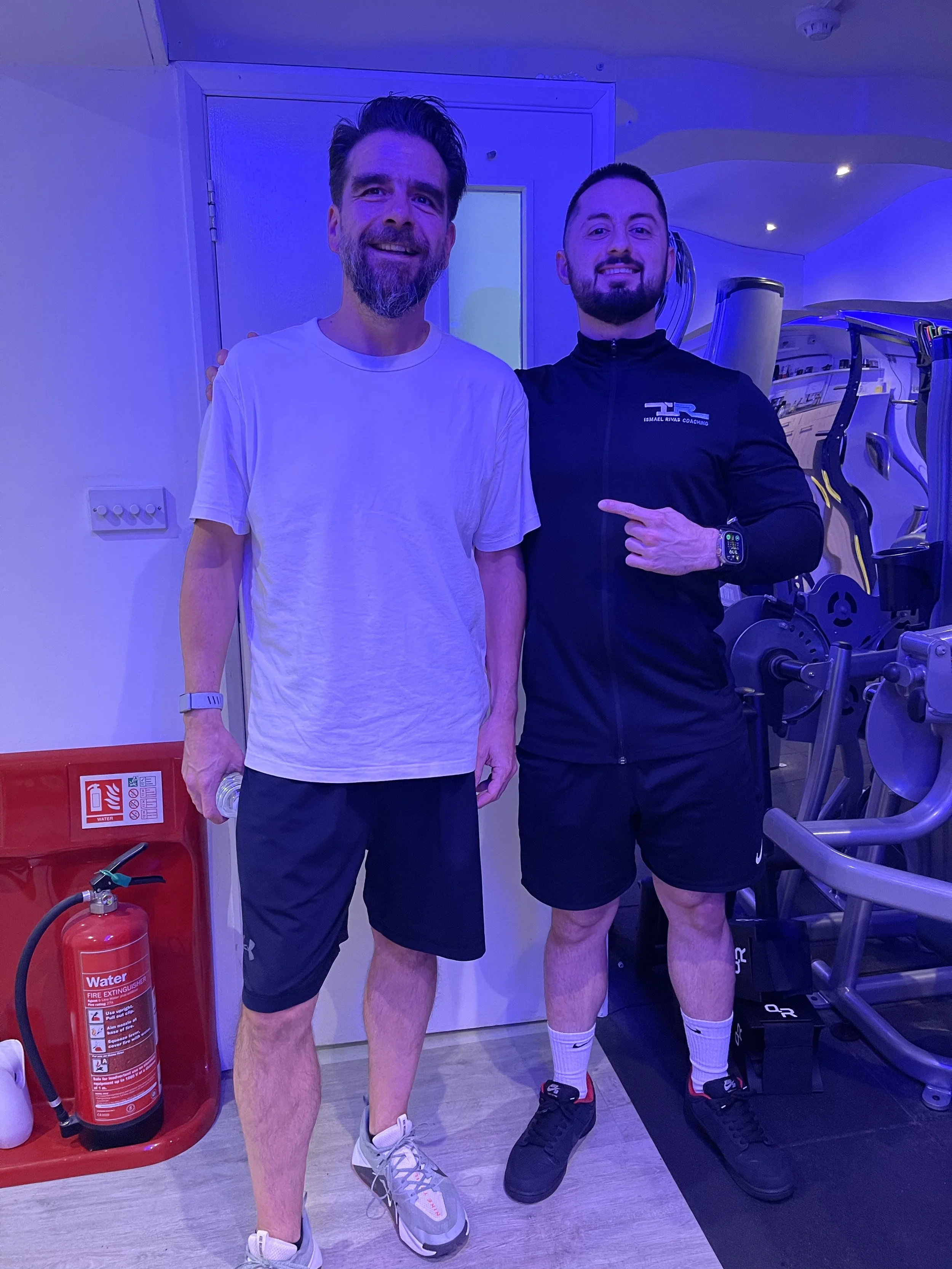 Two men standing in a gym, smiling for the camera. One man is wearing a white T-shirt and black shorts, holding a water bottle. The other man is wearing a black athletic jacket and shorts, pointing at himself. Gym equipment and a fire extinguisher are visible in the background.