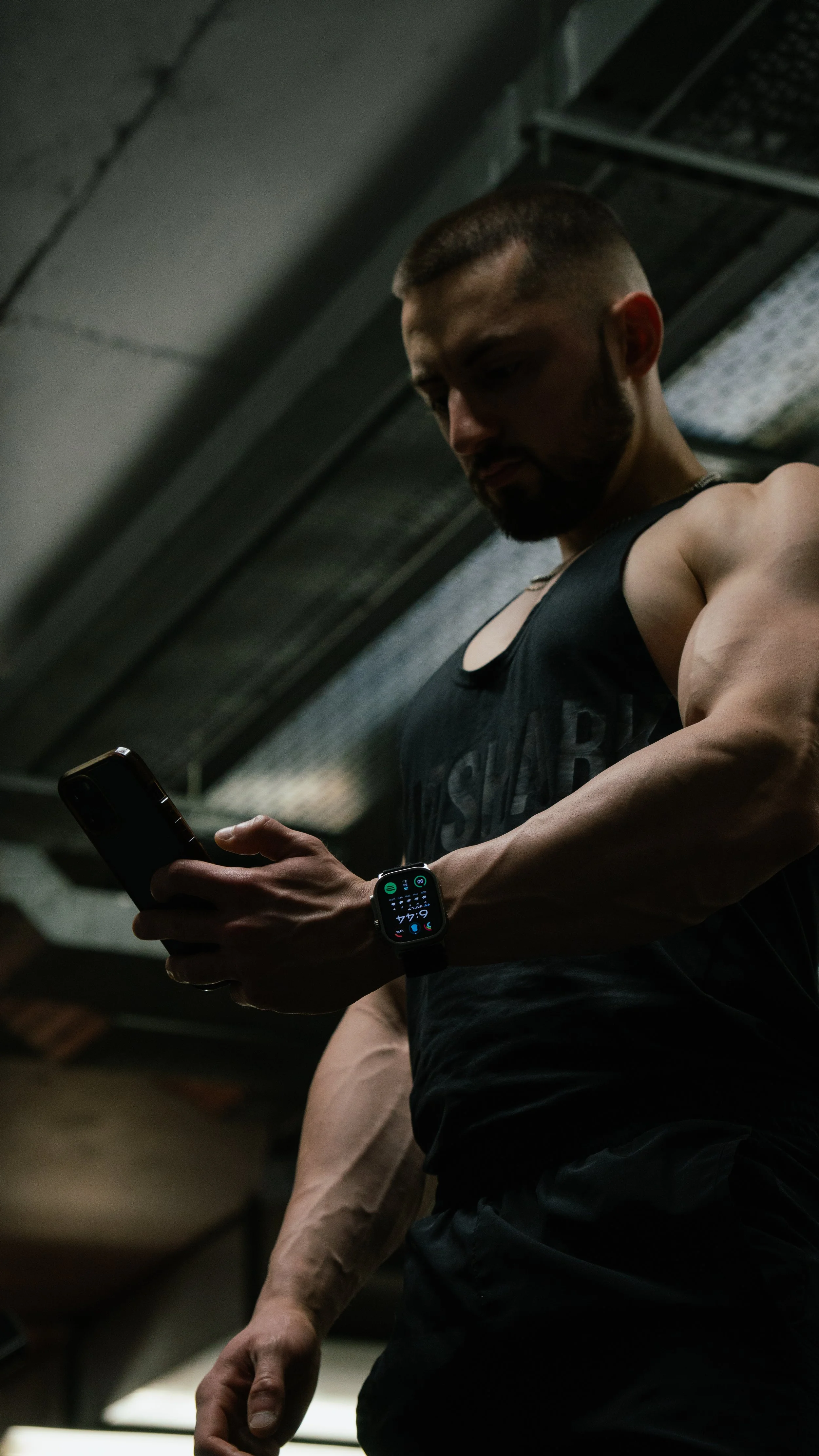 A man in a gym wearing a black sports tank top, looking at his smartphone with a smartwatch on his wrist, under a low ceiling in a fitness setting.