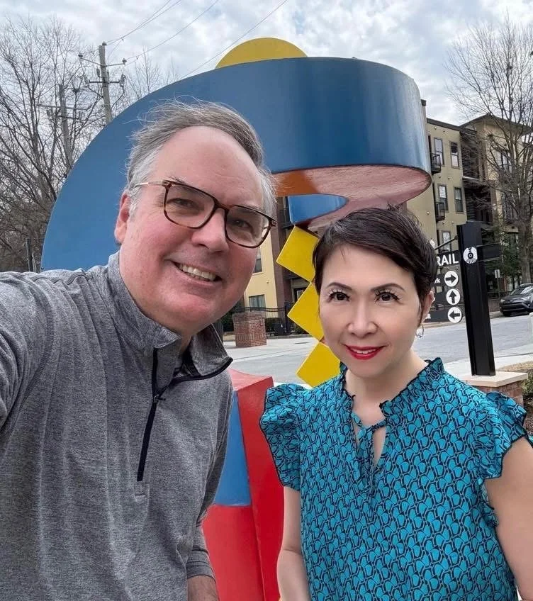 A man and woman taking a selfie outdoors, standing in front of a colorful sculpture with yellow, brown, and blue shapes, and a building and street signs in the background.