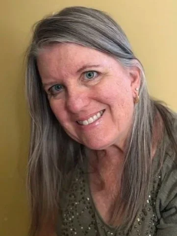 A close-up photo of a smiling woman with long gray hair and blue eyes, wearing a green top with embellishments, against a yellow background.