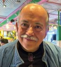 A smiling middle-aged man with a bald head and mustache, wearing a denim jacket indoors at a lively, colorful setting.