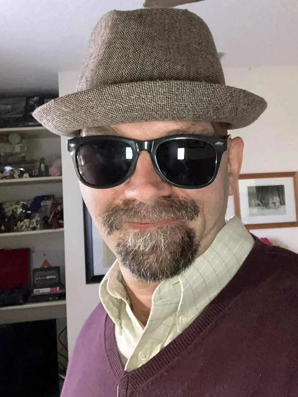 A man wearing a tan hat, black sunglasses, a light yellow collared shirt, and a purple sweater vest.