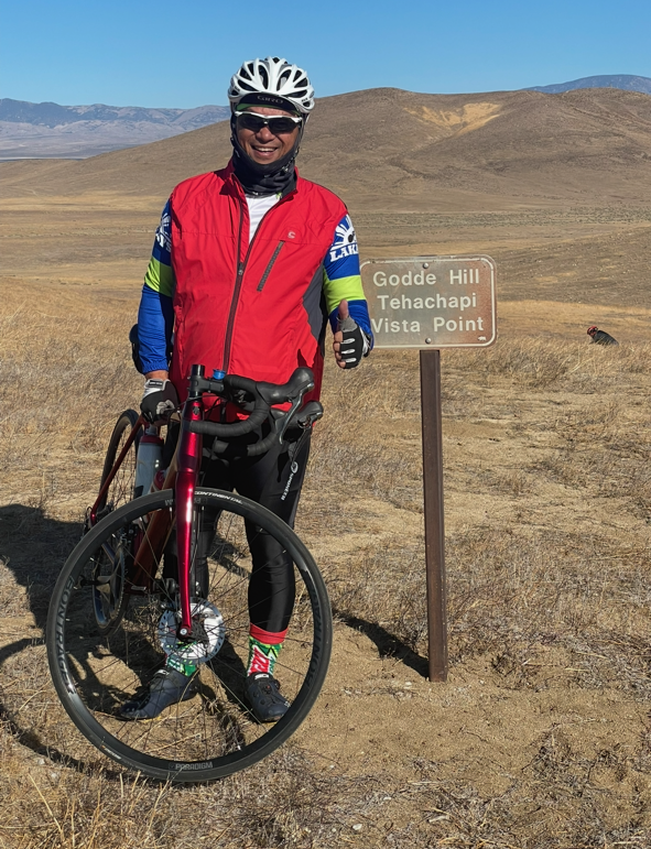 A person in cycling gear and a helmet standing with a bicycle next to a sign that reads 'Godd Hill Tehachapi Vista Point' in a dry, open landscape with hills in the background.