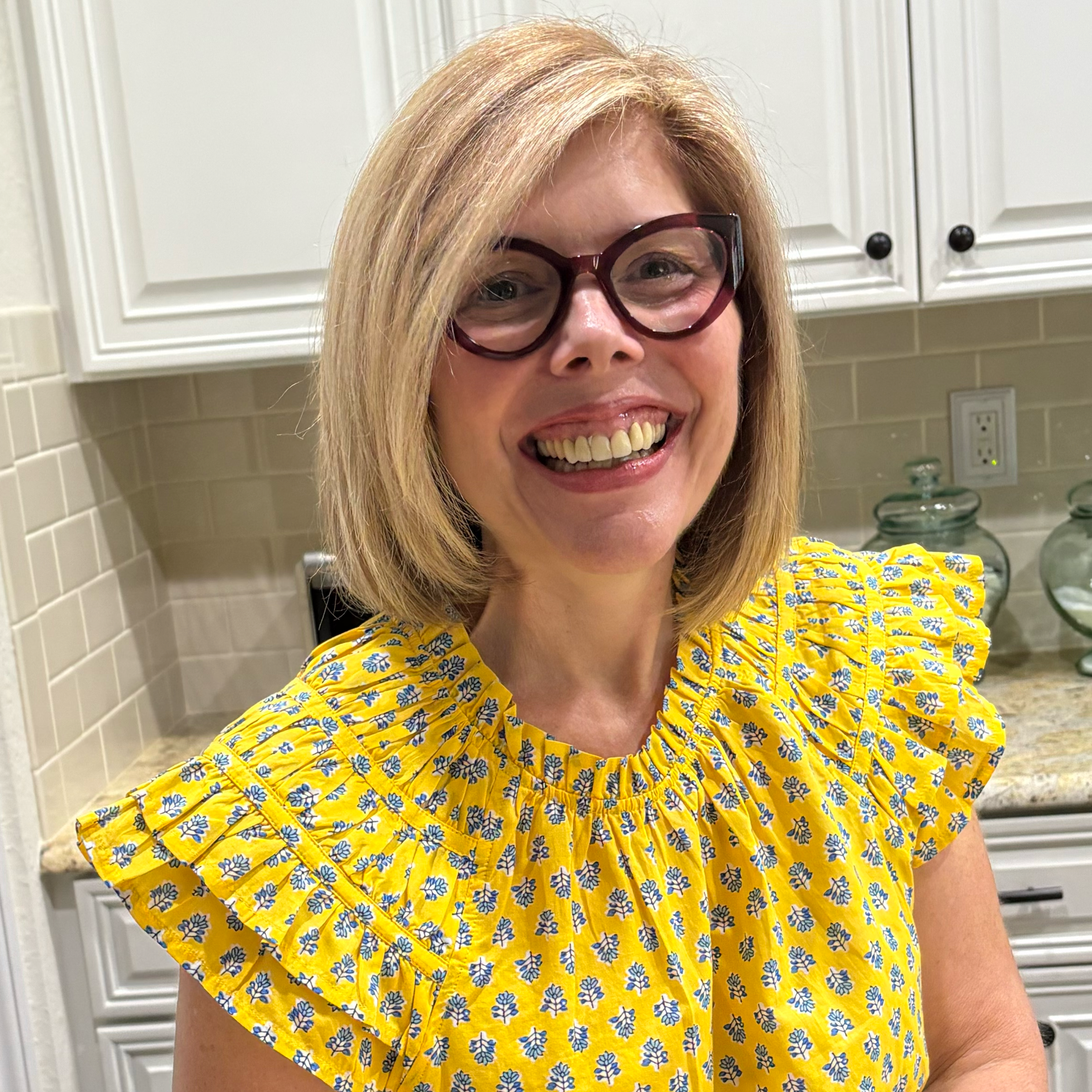 A woman with blonde hair and glasses smiling in a kitchen with white cabinets and a tiled backsplash.