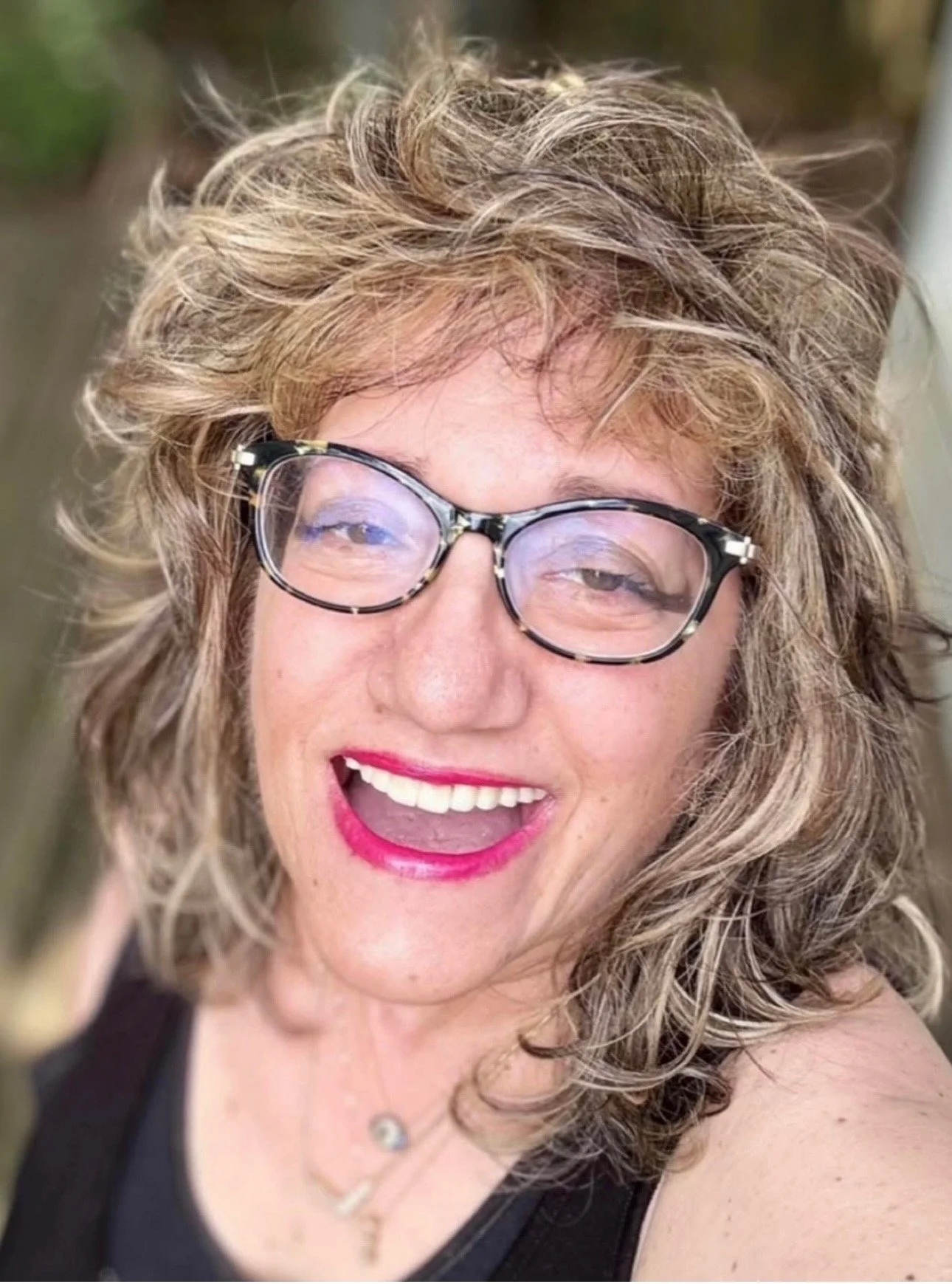 A woman with curly blonde hair wearing glasses and bright pink lipstick, smiling and showing her teeth.