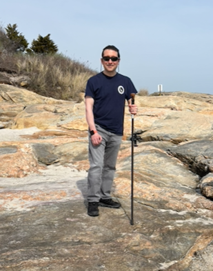 Man standing on rocky shoreline holding a walking stick, wearing a navy T-shirt, gray pants, and sunglasses, with trees and sky in the background.