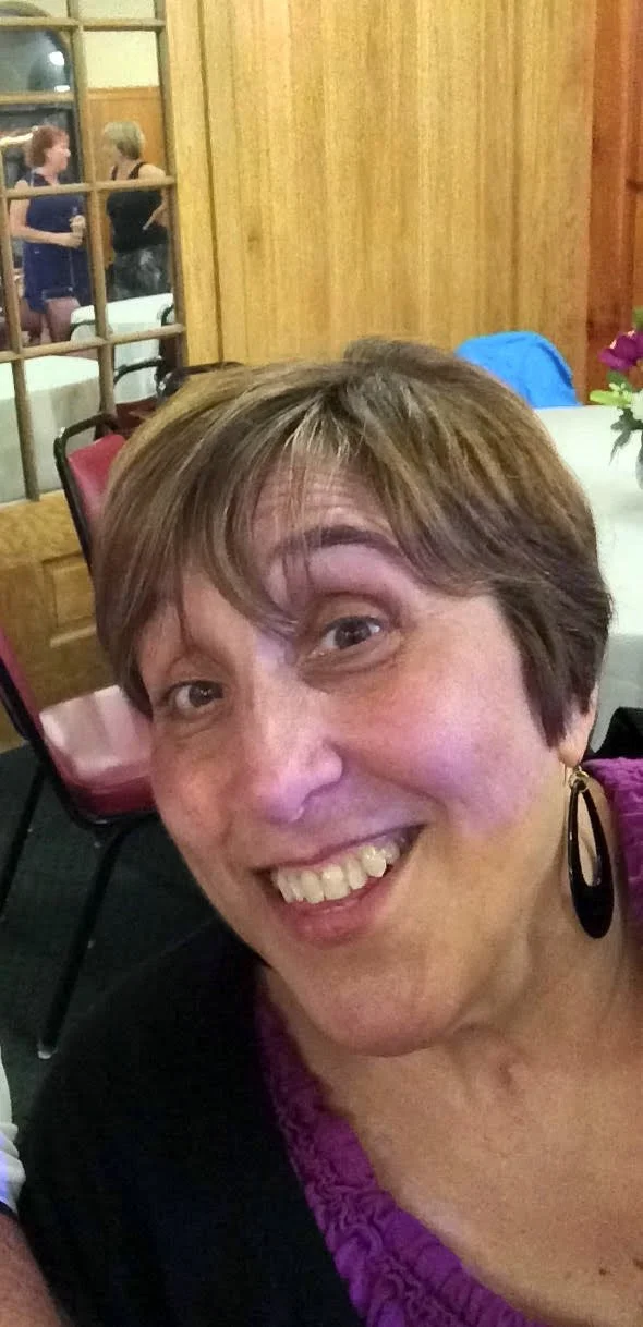Close-up of a woman smiling at a social gathering indoors, with a wooden wall and people talking in the background.