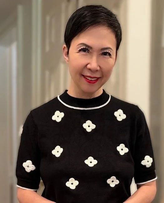 A woman with short black hair, smiling, wearing a black shirt with white floral patterns indoors.