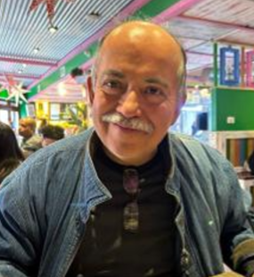 Smiling older man with a mustache and glasses hanging from his shirt, in an arcade or entertainment center with colorful lights and game machines in the background.