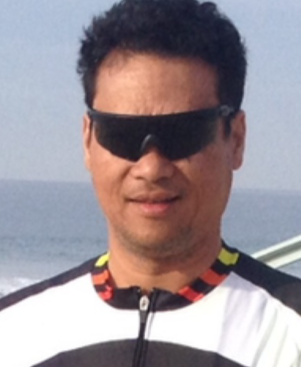 A man wearing sunglasses and a cycling jersey with 'Pedal Power' written on it, standing outdoors near water.