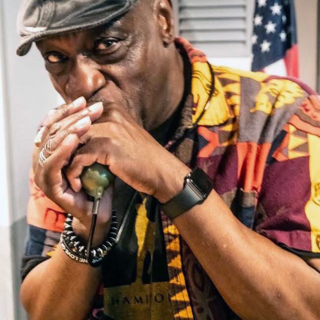 Man in colorful patterned shirt holding a telephone receiver to his mouth, with black watch and bracelets, in front of an American flag.