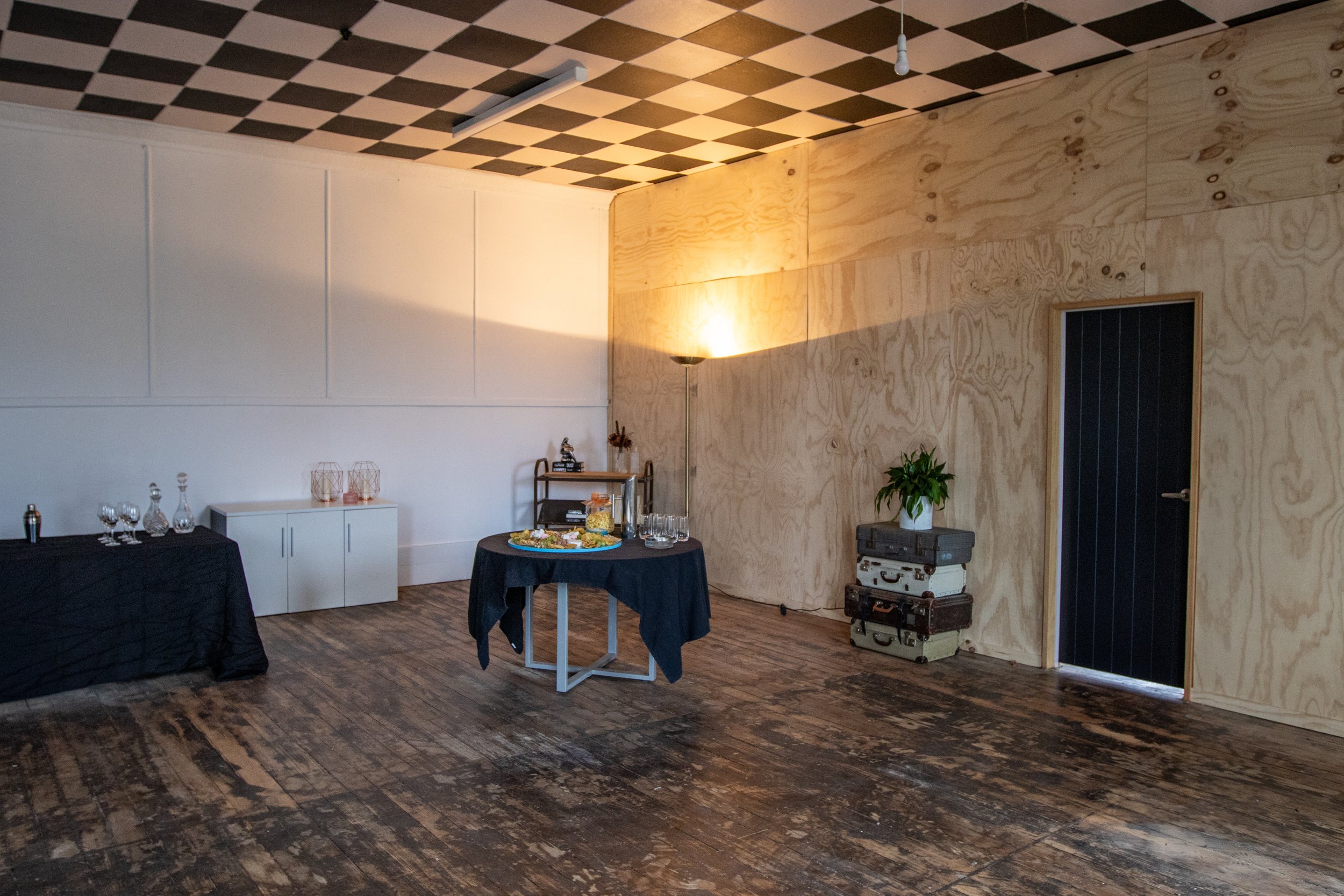 Interior of a room with checkered ceiling, plywood wall, and a dark hardwood floor, featuring a small table with a bowl of snacks and a potted plant, a black-draped table with glassware, and a corner with stacked vintage suitcases and a lamp.