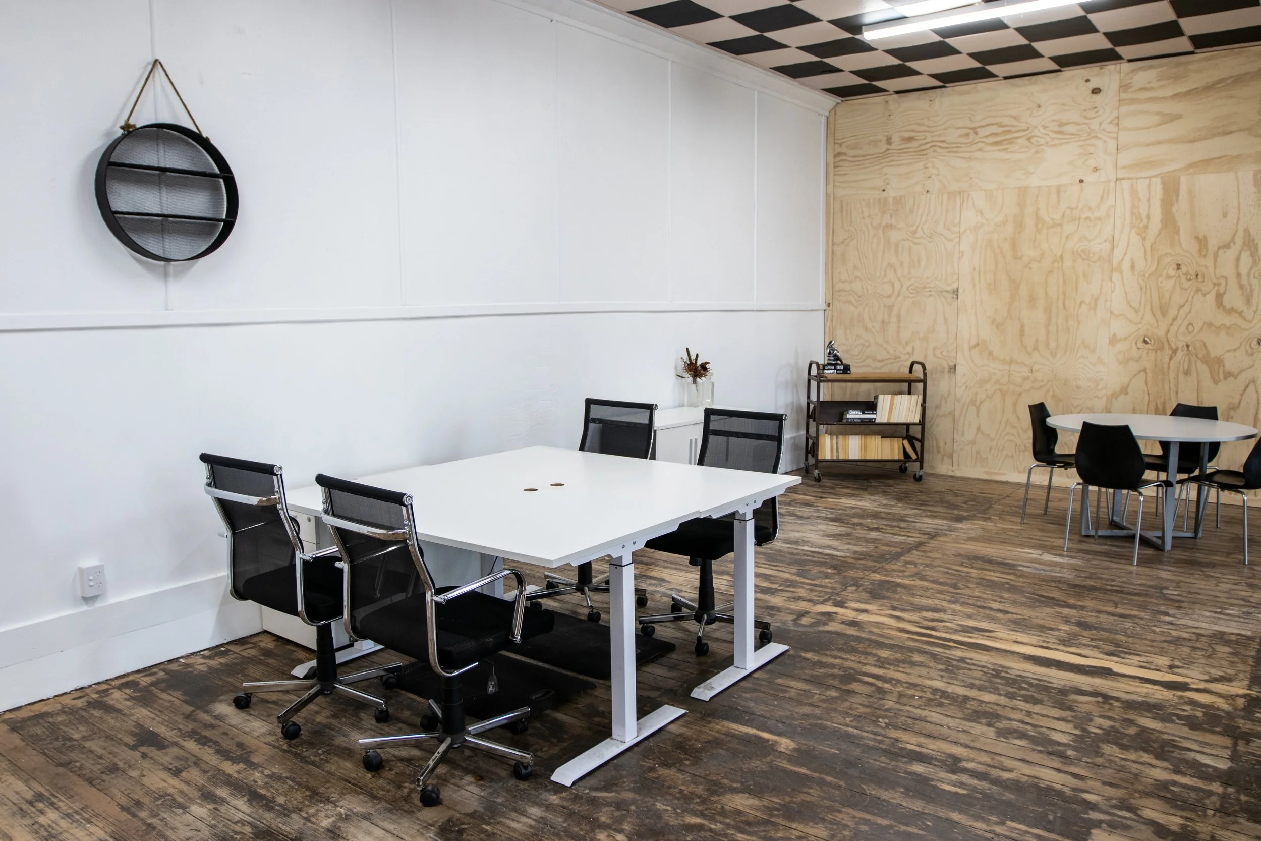 A modern coworking space with white walls, wooden flooring, and a variety of seating arrangements including black chairs around a white table and a small round table with black chairs. There is a black wall clock, a small wooden shelf, and decorative items like a glass jar with flowers. The ceiling has black and white checkered tiles.