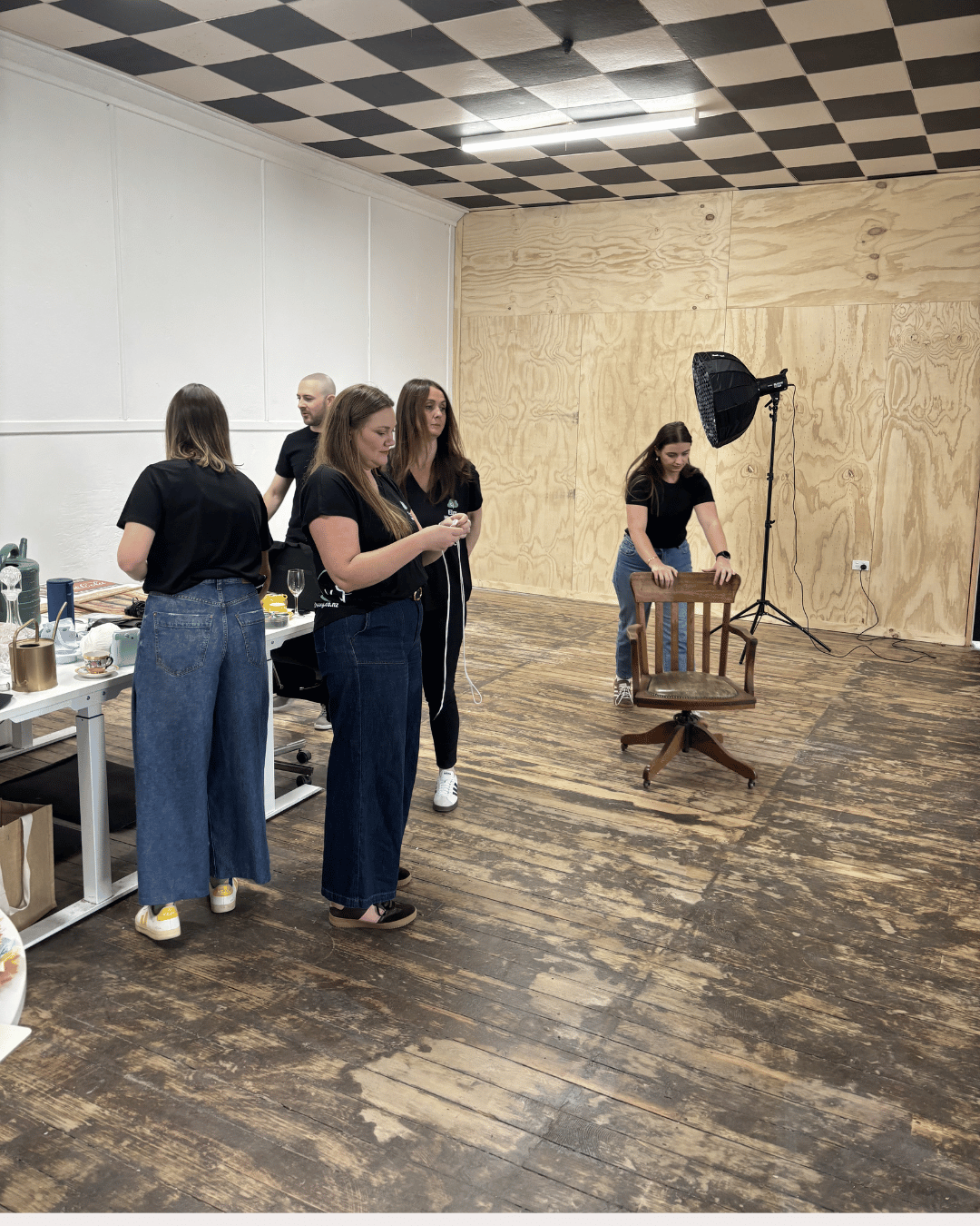 Five women in black shirts and jeans preparing in an indoor studio with a wooden floor, a large softbox light, and a chair.
