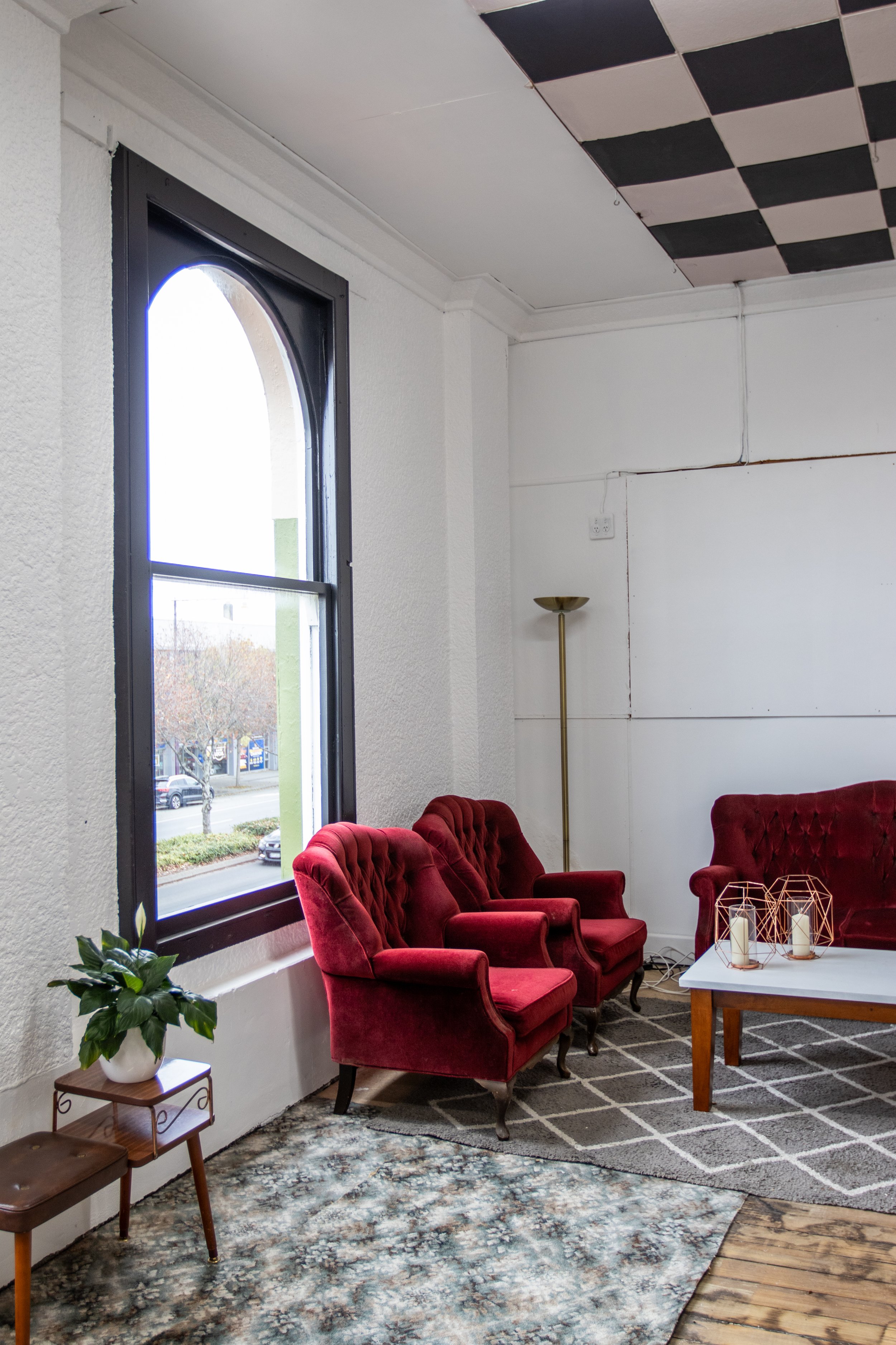 A cozy indoor sitting area with four red velvet armchairs surrounding a white coffee table with candles and decorative wire lanterns. There is a dark patterned rug, a tall floor lamp, and a small side table with potted plant. A large window with black trim lets in natural light. The ceiling has a black and white checkered pattern.