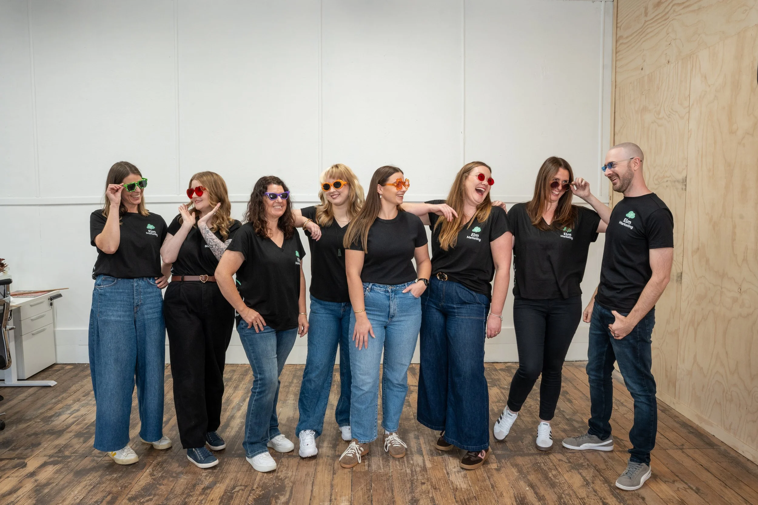A group of seven people, six women and one man, standing indoors on a wooden floor with a plain wall behind them, wearing matching black t-shirts with a green logo and sunglasses in different bright colors, smiling and posing together.