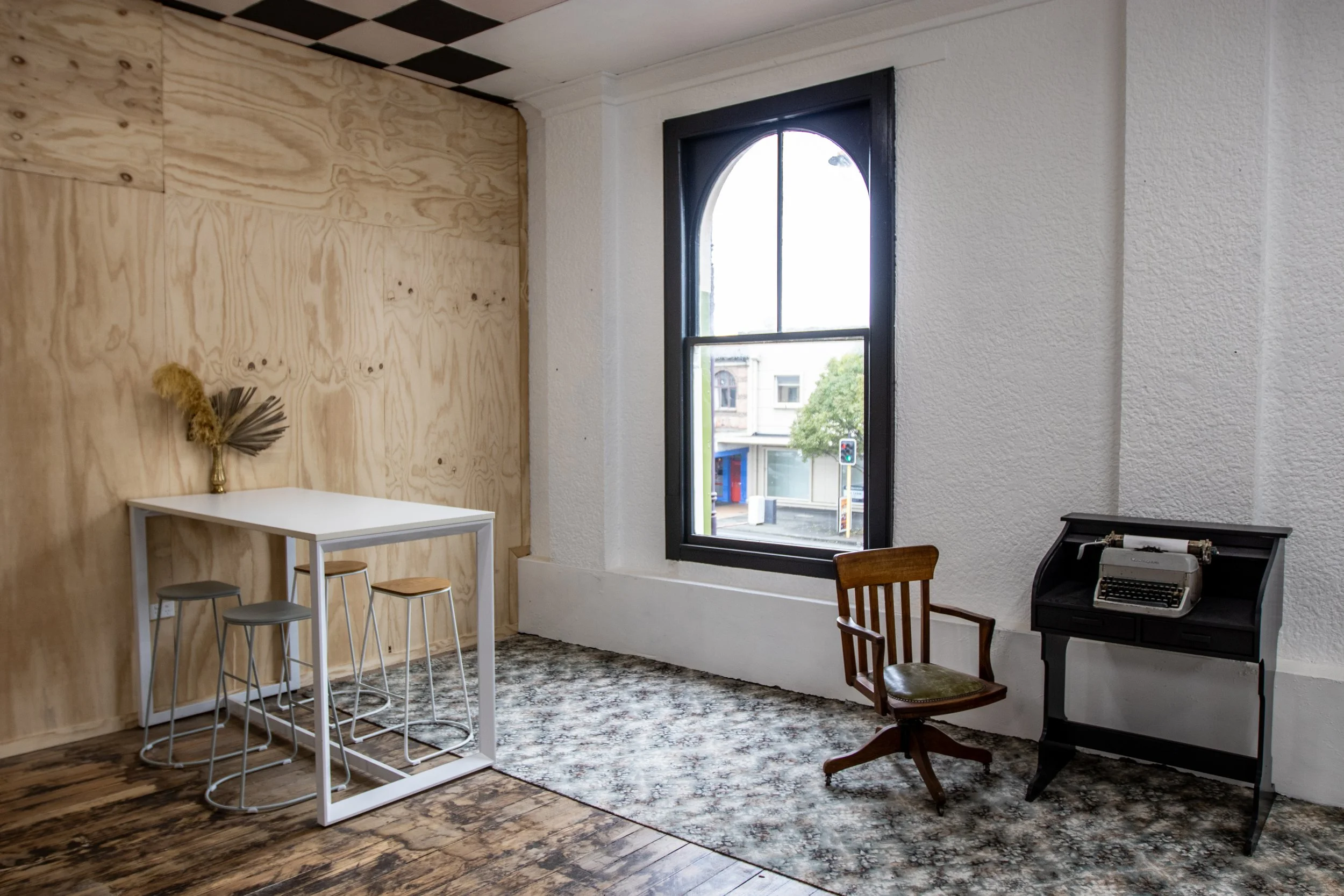 Interior of a room with a wooden accent wall, a high table with three stools, a large window showing a city street, a wooden chair, and an old-fashioned typewriter on a black stand.
