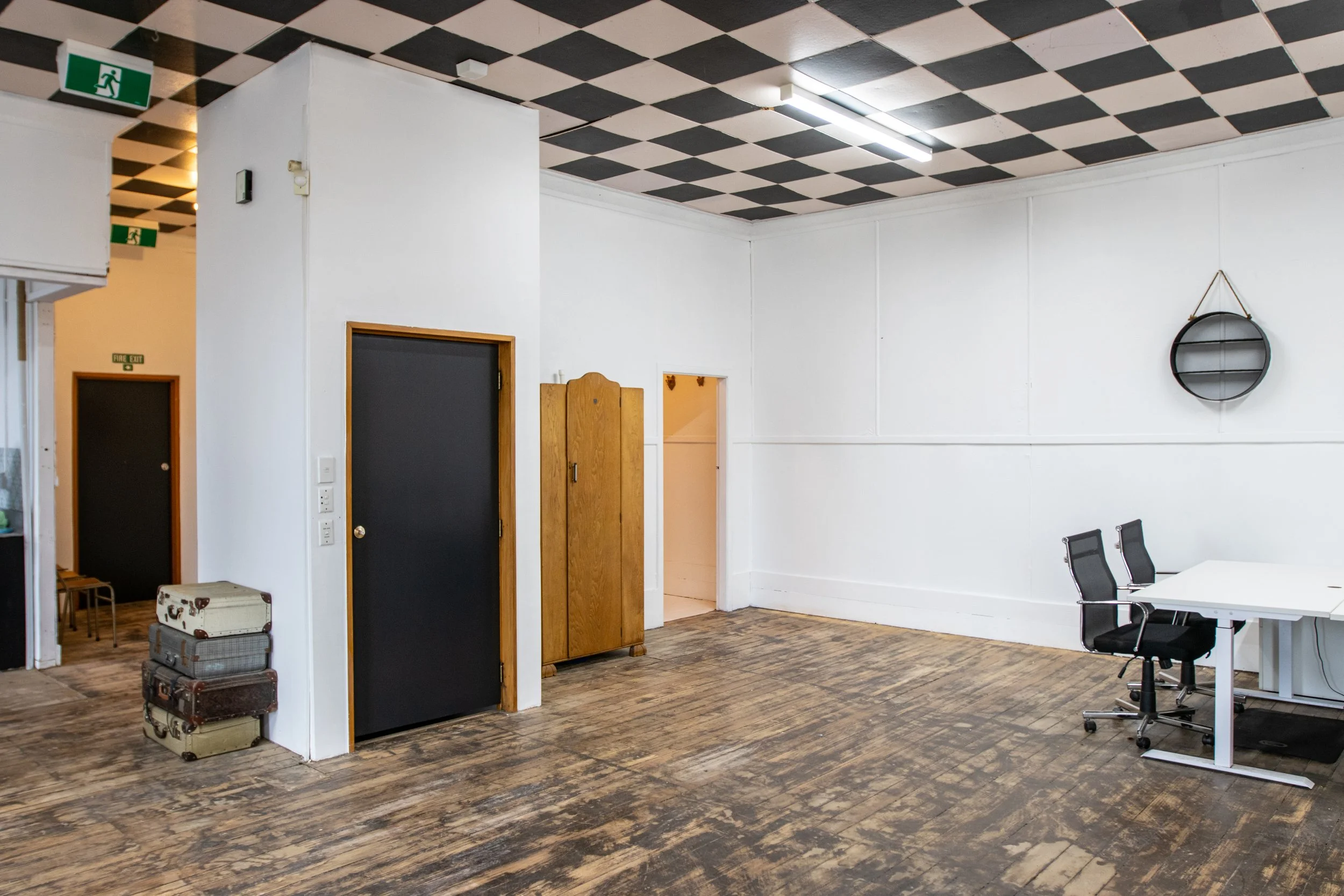 Empty room with white walls, dark checkered ceiling, wooden floor, stacked suitcases, dark door, round wall mirror, and white table with black office chairs.
