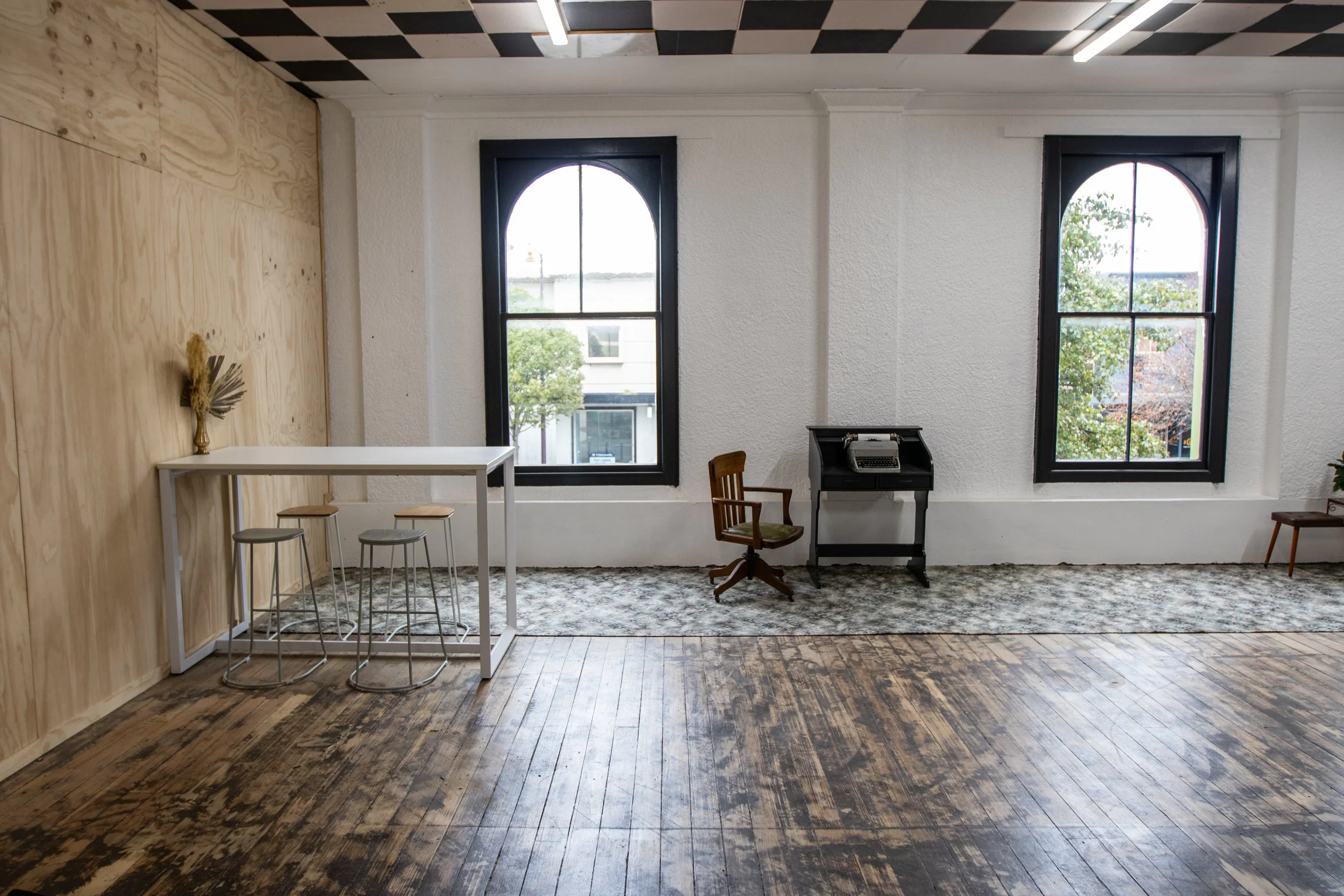 Interior view of a room with two large arched windows, white textured walls, dark wood floor, a small black desk with a typewriter, a wooden chair, a white table with four stools, and a decorative vase on the wall assembly.