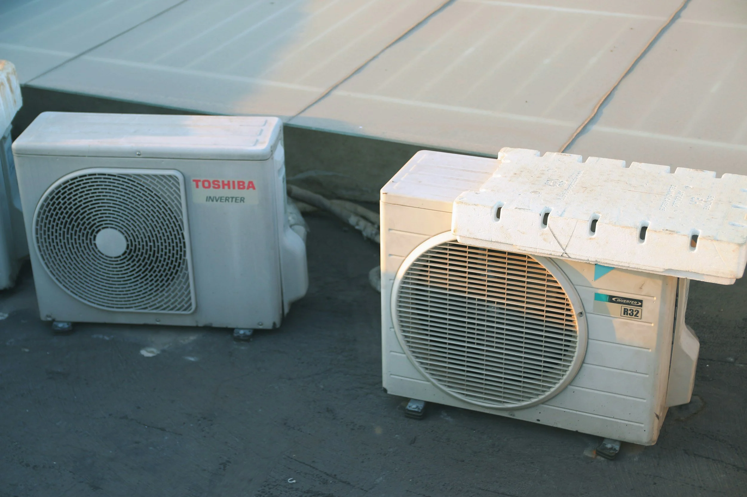 Two outdoor air conditioning units on a rooftop, one branded Toshiba and the other appears to be an older model, sitting on a flat surface with a corrugated metal roof above.