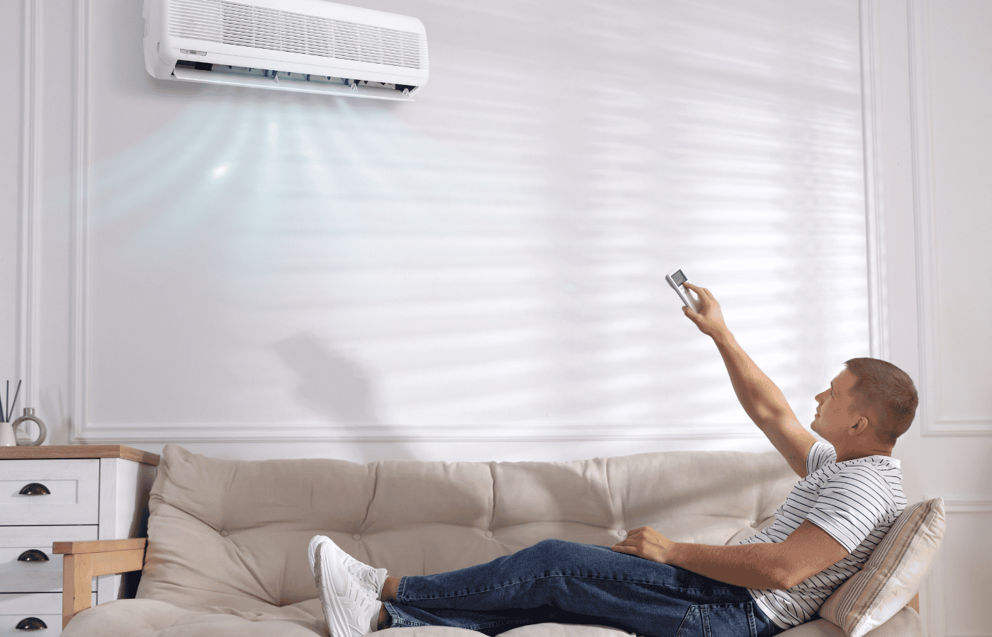 A young man sitting on a beige couch in a living room, holding a remote control towards an air conditioning unit mounted on the wall, with sunlight creating pattern shadows through blinds on the wall.