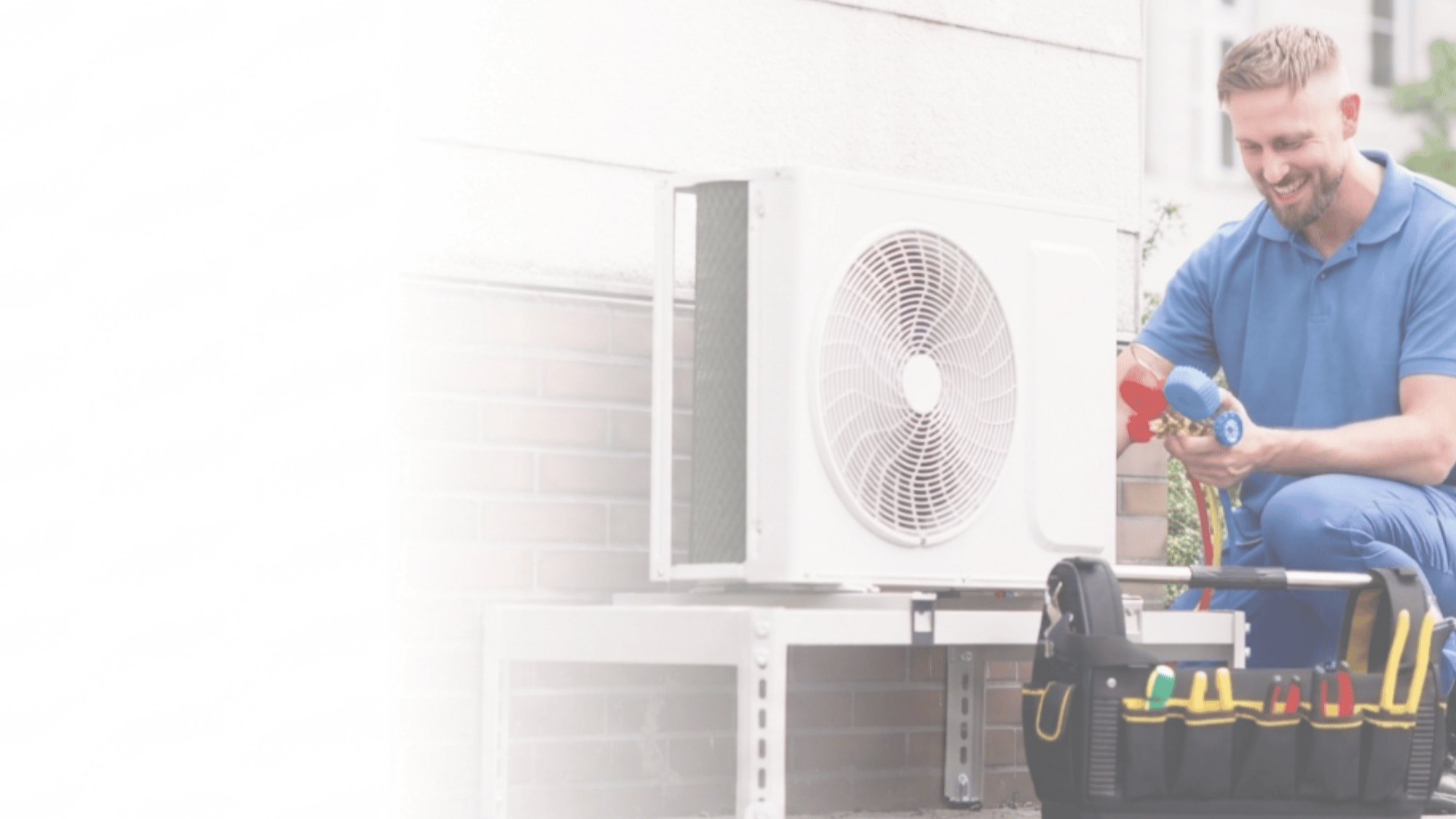 A man in a blue shirt working on an air conditioning unit outside a building, holding tools and a garden hose.