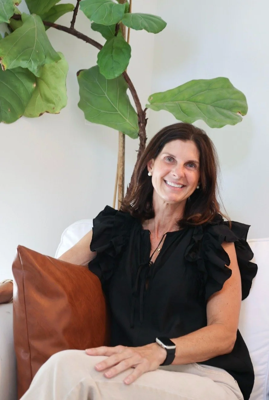 A woman with shoulder-length brown hair and a black ruffled top sitting on a white sofa with a tan leather pillow, smiling, in front of a large indoor fig tree with big green leaves.