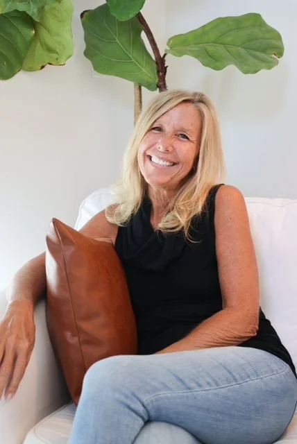 A smiling woman with blonde hair sitting on a white sofa, holding a brown pillow, with a large green plant behind her.