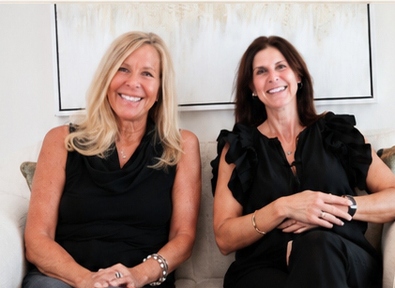 Two women sitting on a light-colored couch, smiling, with a white wall and framed art behind them. The woman on the left has long blonde hair, and the woman on the right has short brown hair. Both are wearing black tops.
