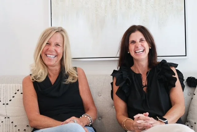 Two women sitting on a light-colored couch, laughing and smiling, with a whiteboard in the background.