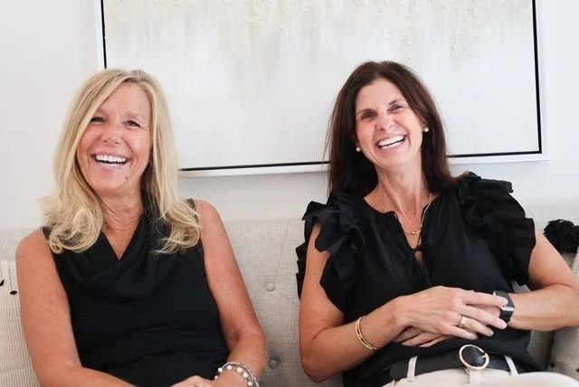 Two women sitting on a beige couch, smiling and laughing, in a room with a white wall and a large screen behind them.