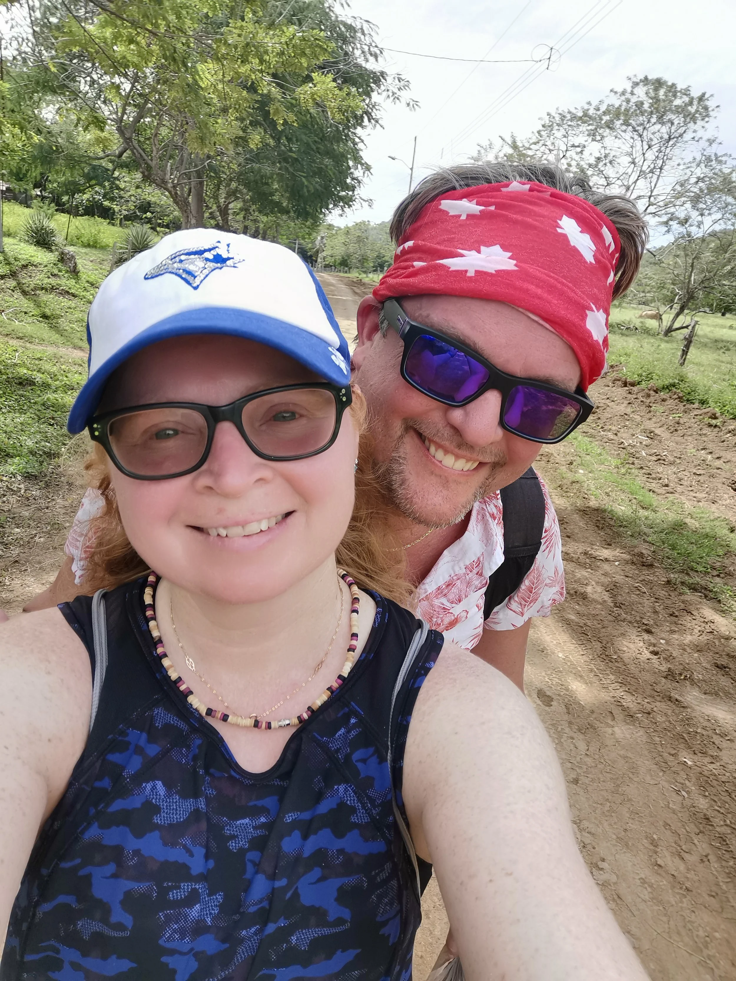 Close-up of a smiling man and woman outdoors on a dirt trail. The woman is wearing glasses, a blue camouflage-patterned top, a colorful beaded necklace, and a white and blue cap. The man has a red bandana with white maple leaves, sunglasses, and a white shirt with a red pattern. The background features trees, grass, and power lines.