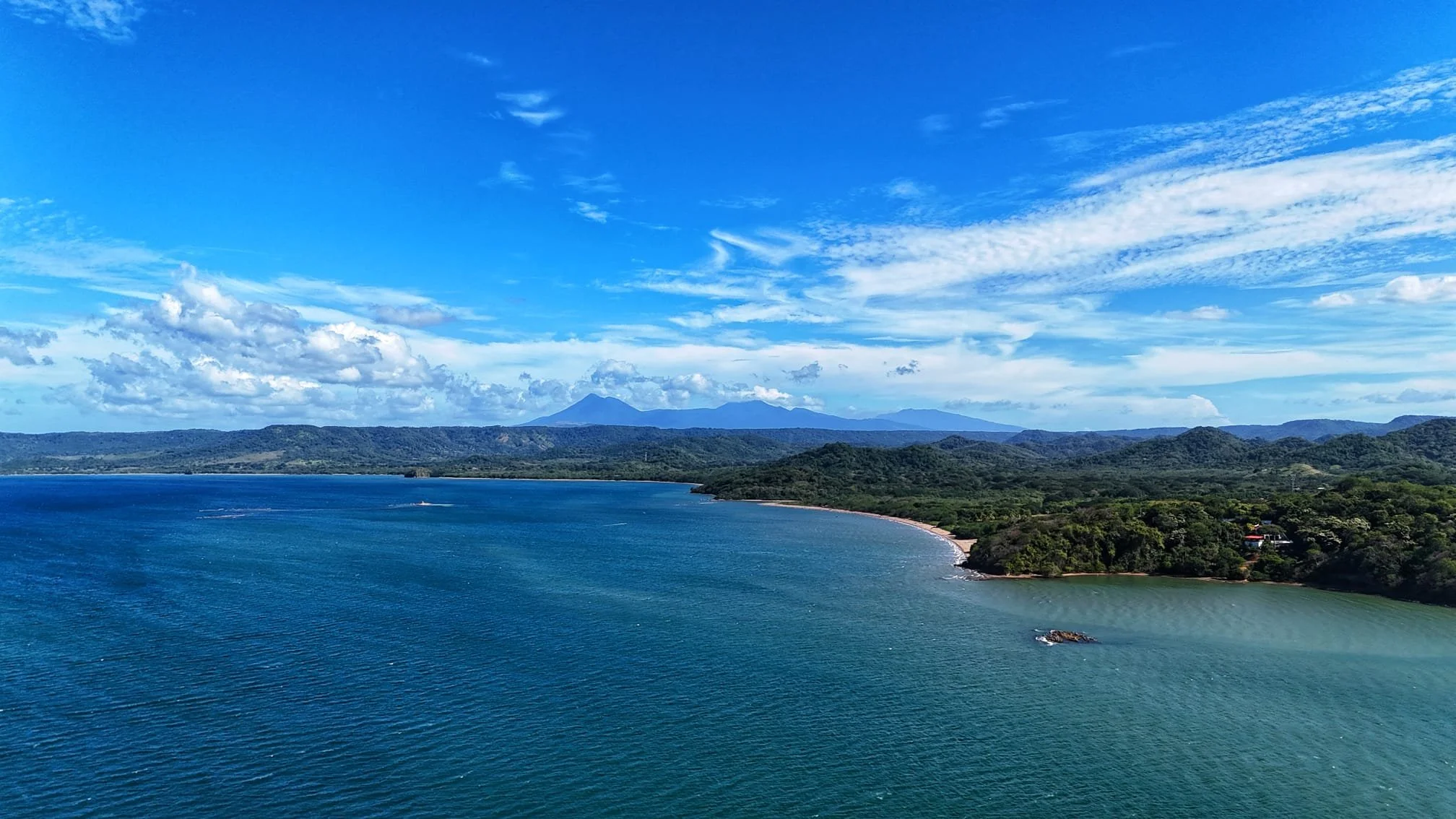 A scenic view of a large body of water with a shoreline, lush green hills, and distant mountains under a mostly clear blue sky with scattered clouds.