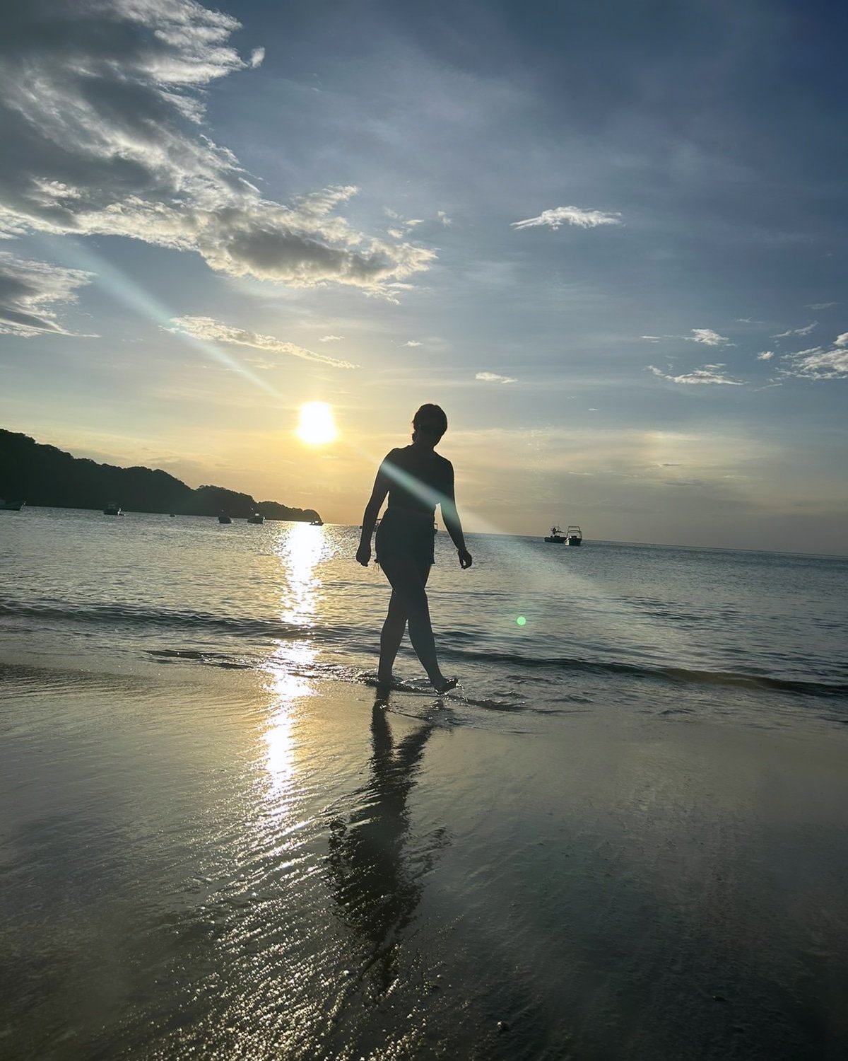 Person walking along the shoreline at sunset with boats in the water and a cloudy sky.