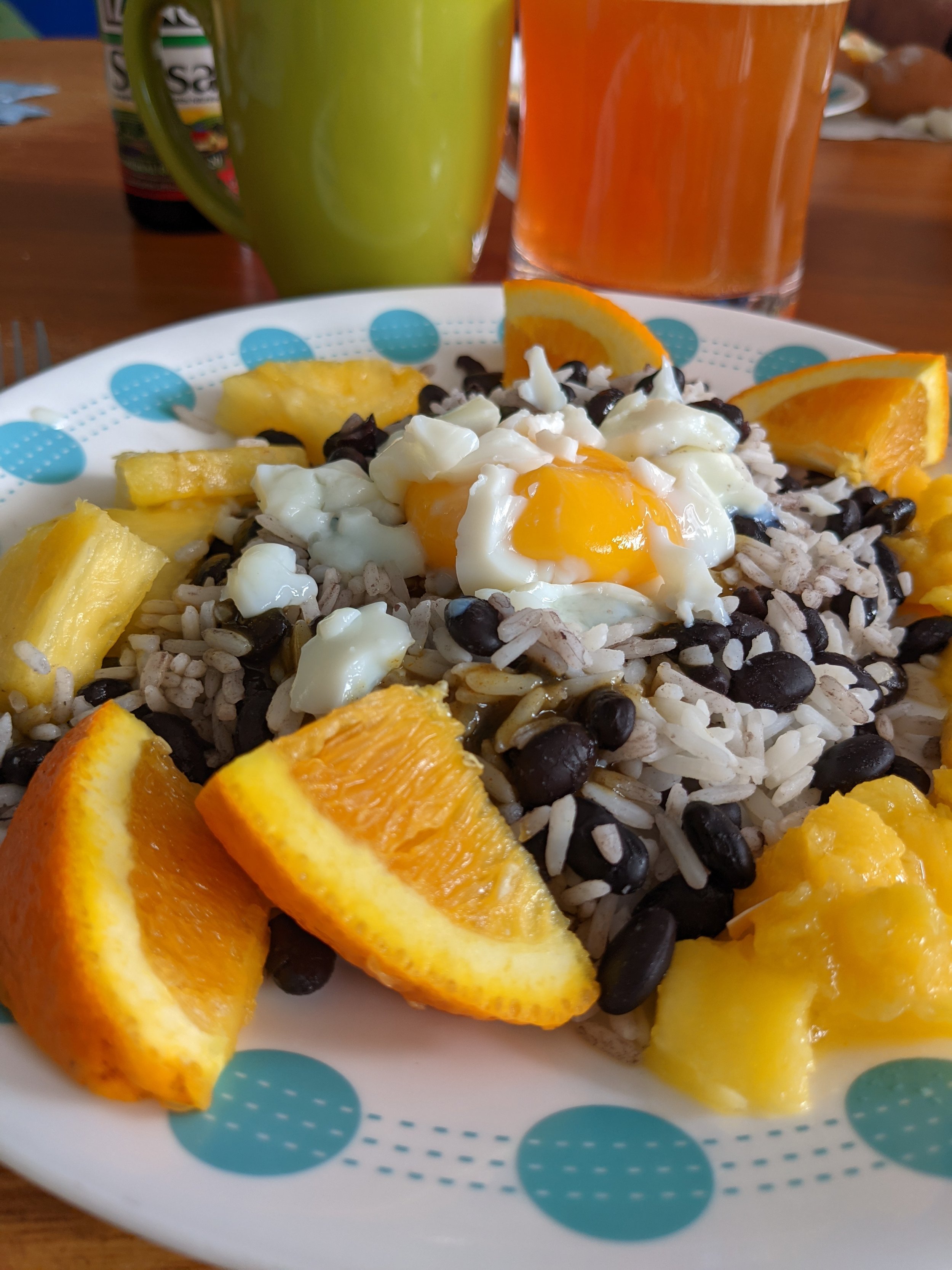 A breakfast plate with rice, black beans, a soft-boiled egg with a runny yolk, orange slices, and pineapple chunks. In the background, there's a green mug and an orange beverage in a glass.