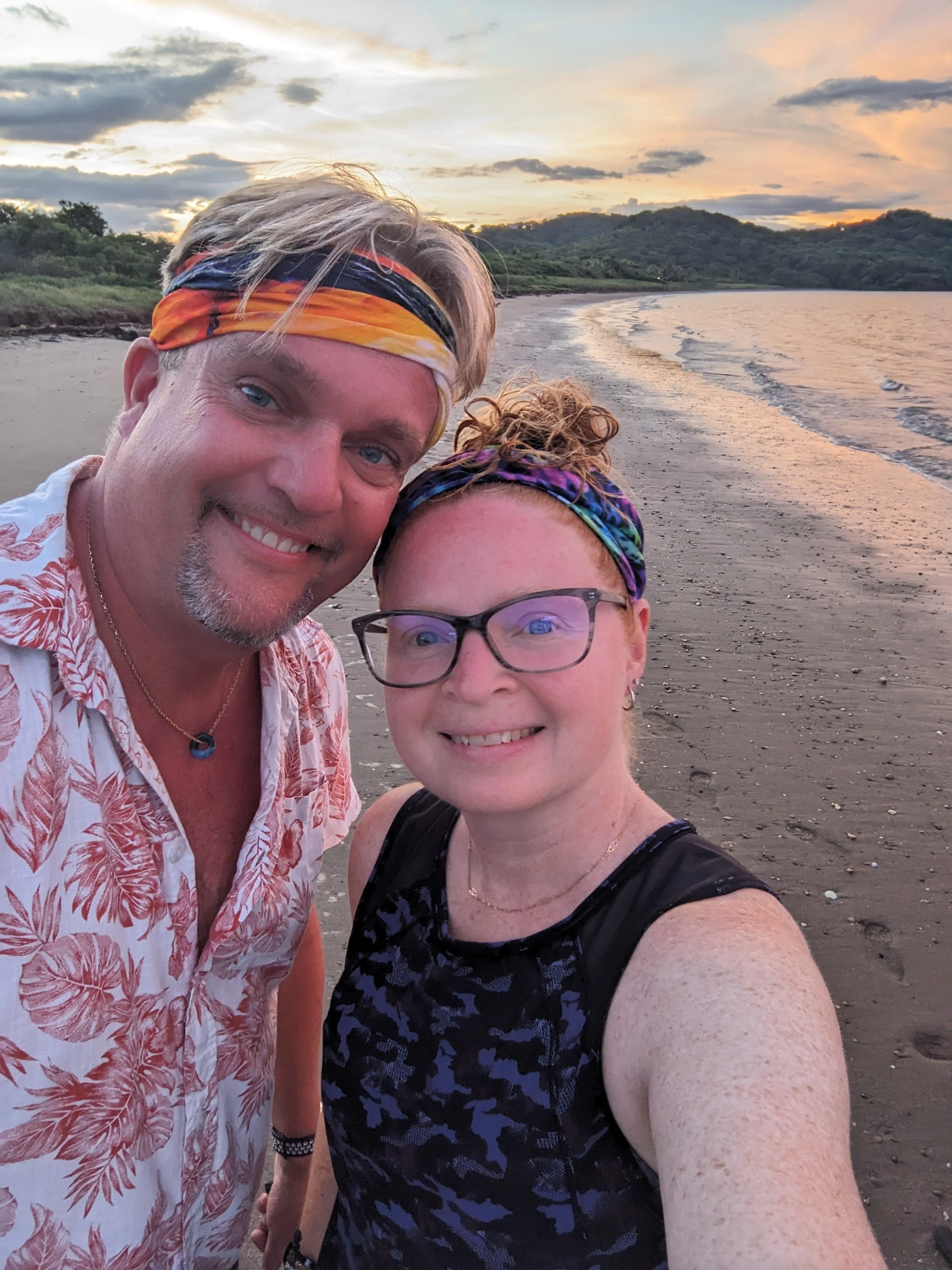 A couple taking a selfie on a beach at sunset, with green hills in the background and cloudy sky.
