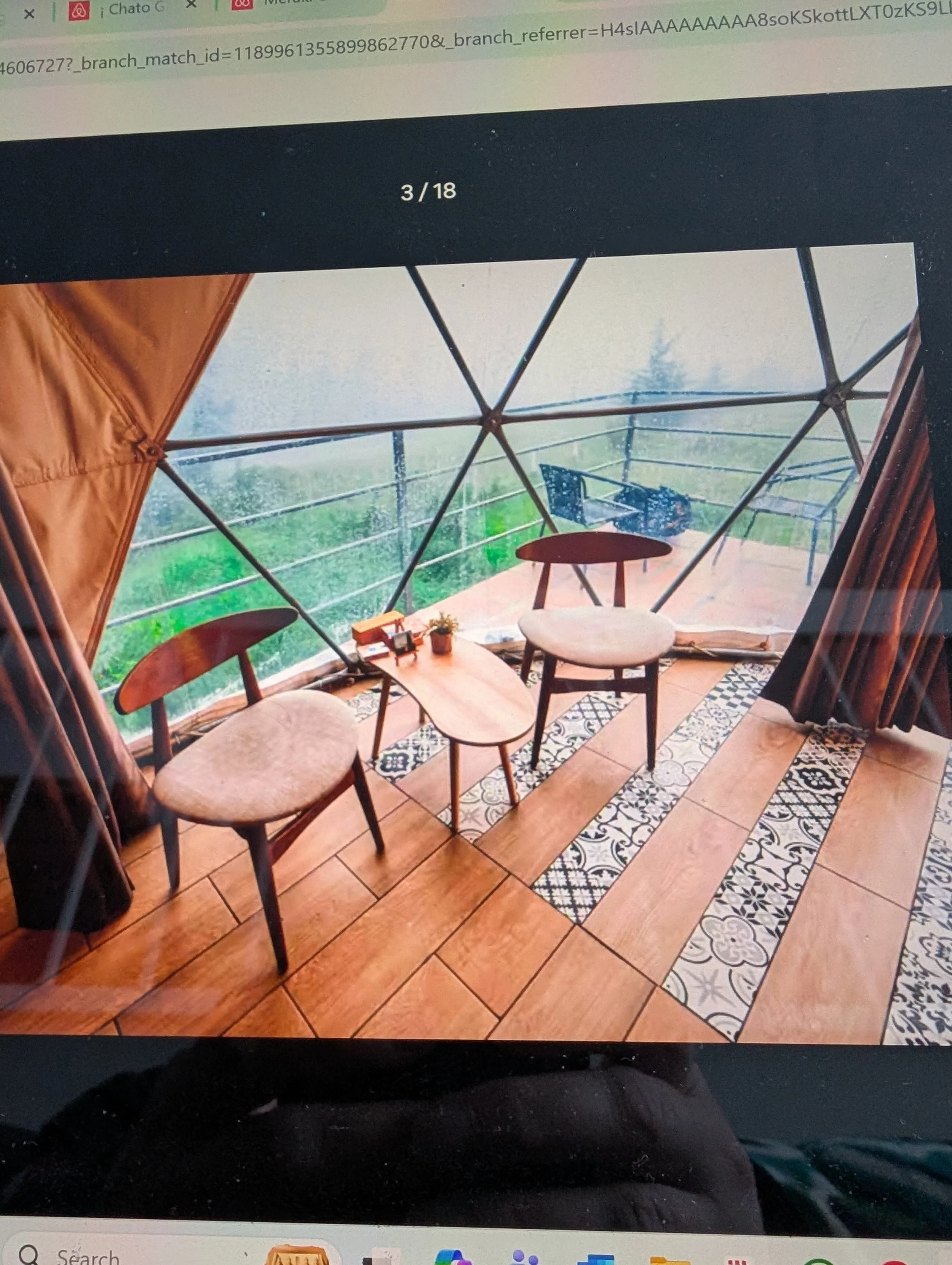 Inside a geodesic dome with seating, tables, and decorated floors, overlooking a grassy landscape and trees on a rainy day.