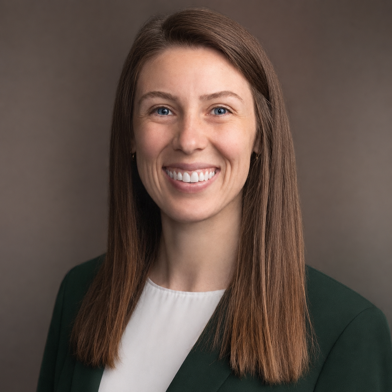 A professional woman with long, straight brown hair, blue eyes, wearing a white top and dark blazer, smiling against a neutral gray background.