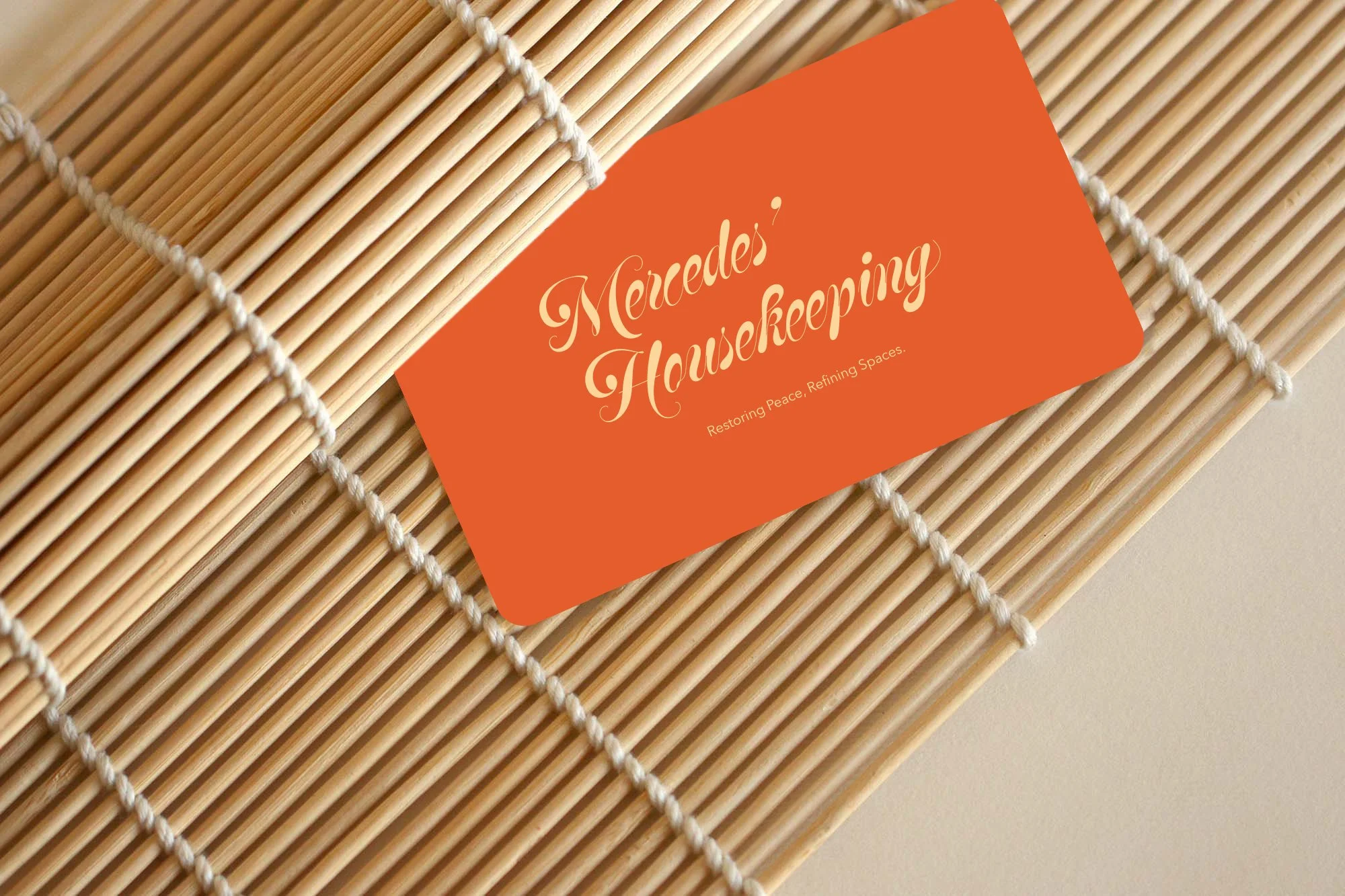 A business card for Mercedes' Housekeeping resting on a wooden slat surface. The card is orange with cream-colored script text.