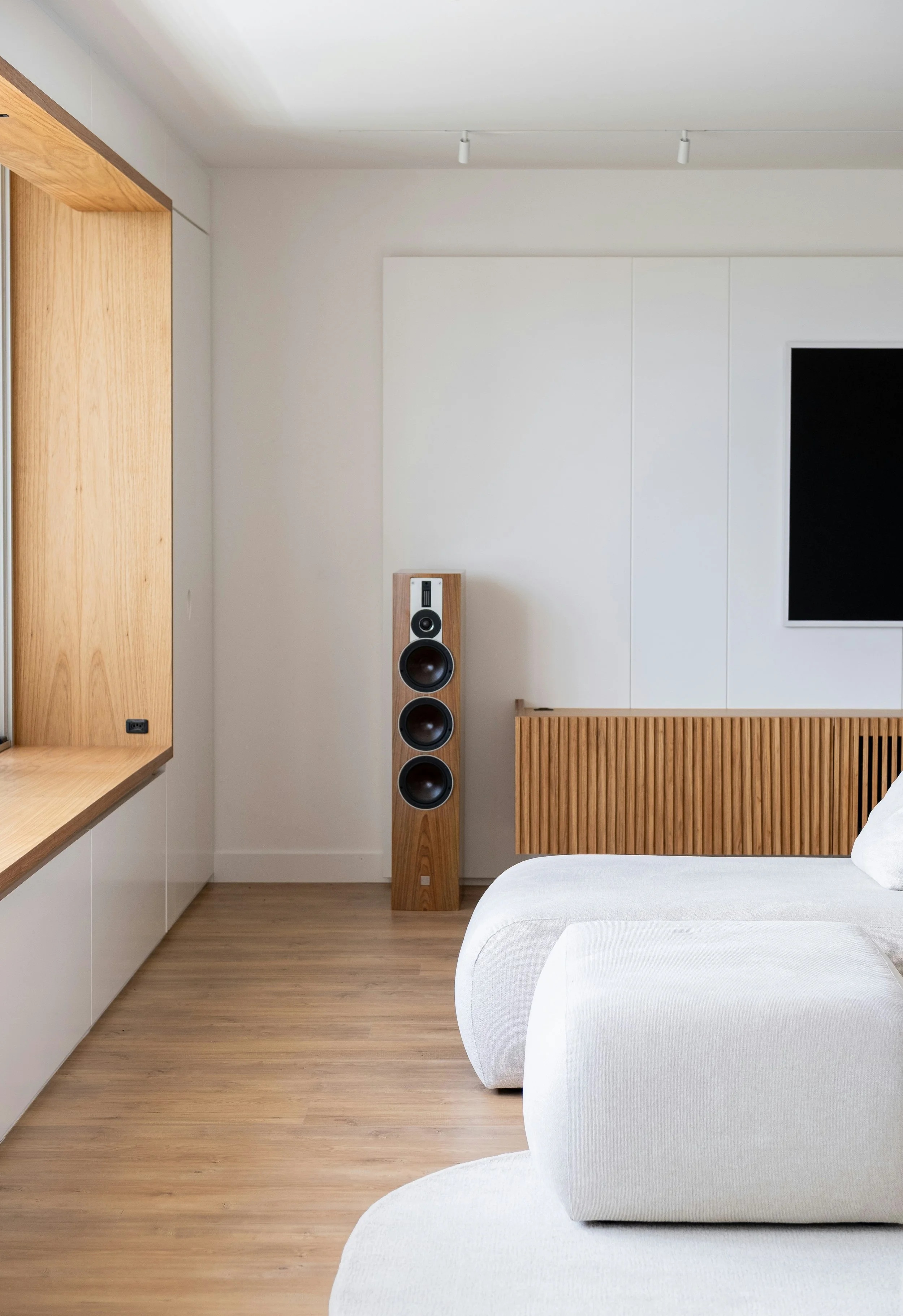A modern bedroom with white walls, light wood flooring, a white upholstered bed, and a tall wooden speaker on the floor, with a mounted flat-screen TV on the wall.