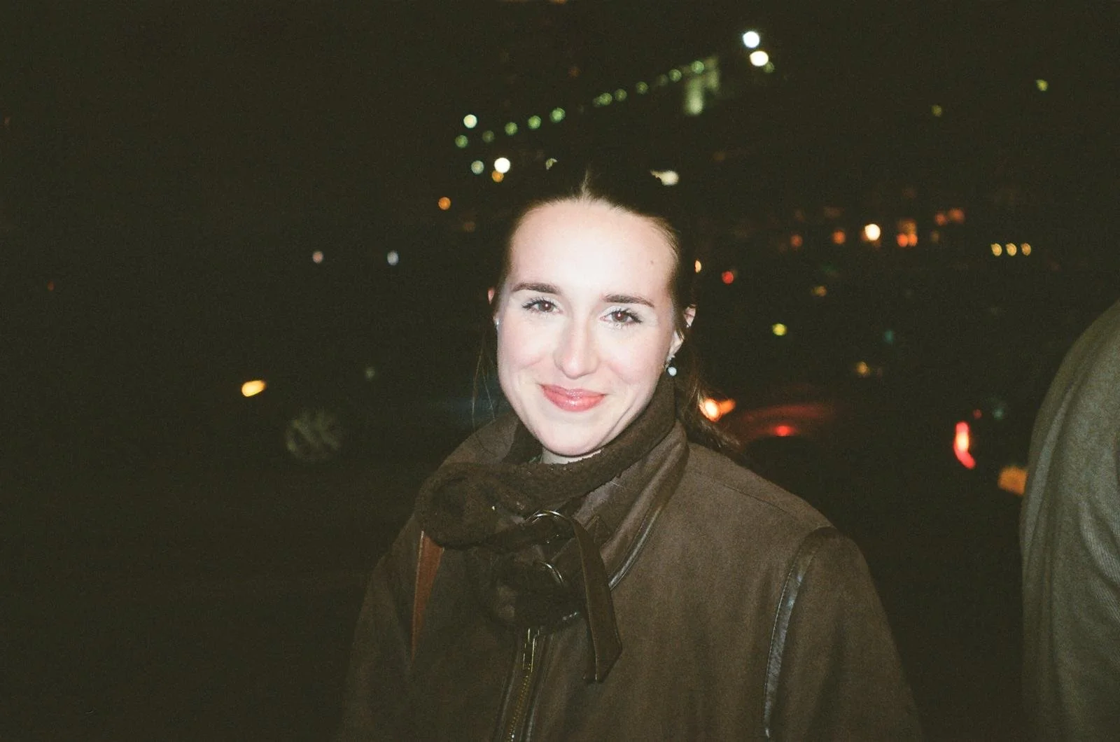 A woman with light skin and dark hair tied back, smiling at night. She is wearing a dark brown jacket and a scarf. The background features blurry city lights.