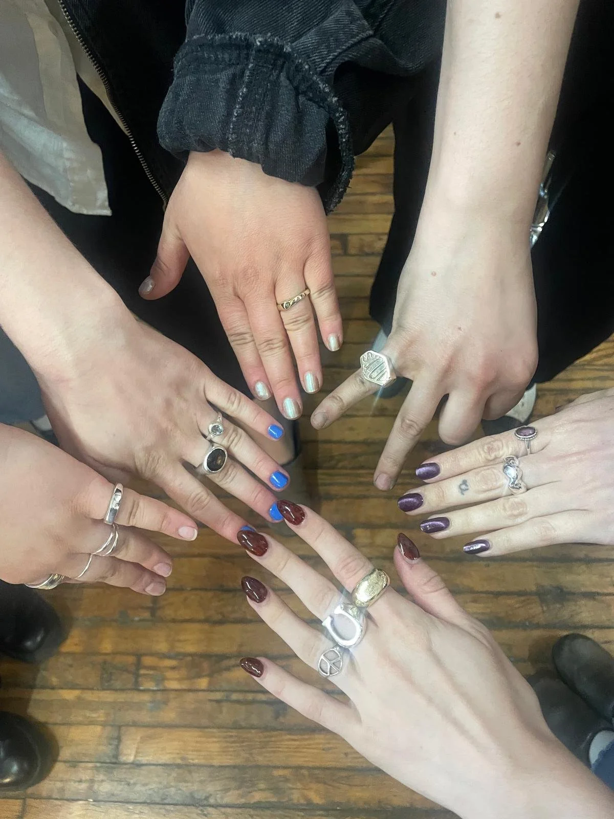 Multiple hands with rings and painted nails, positioned in a star shape on a wooden floor.
