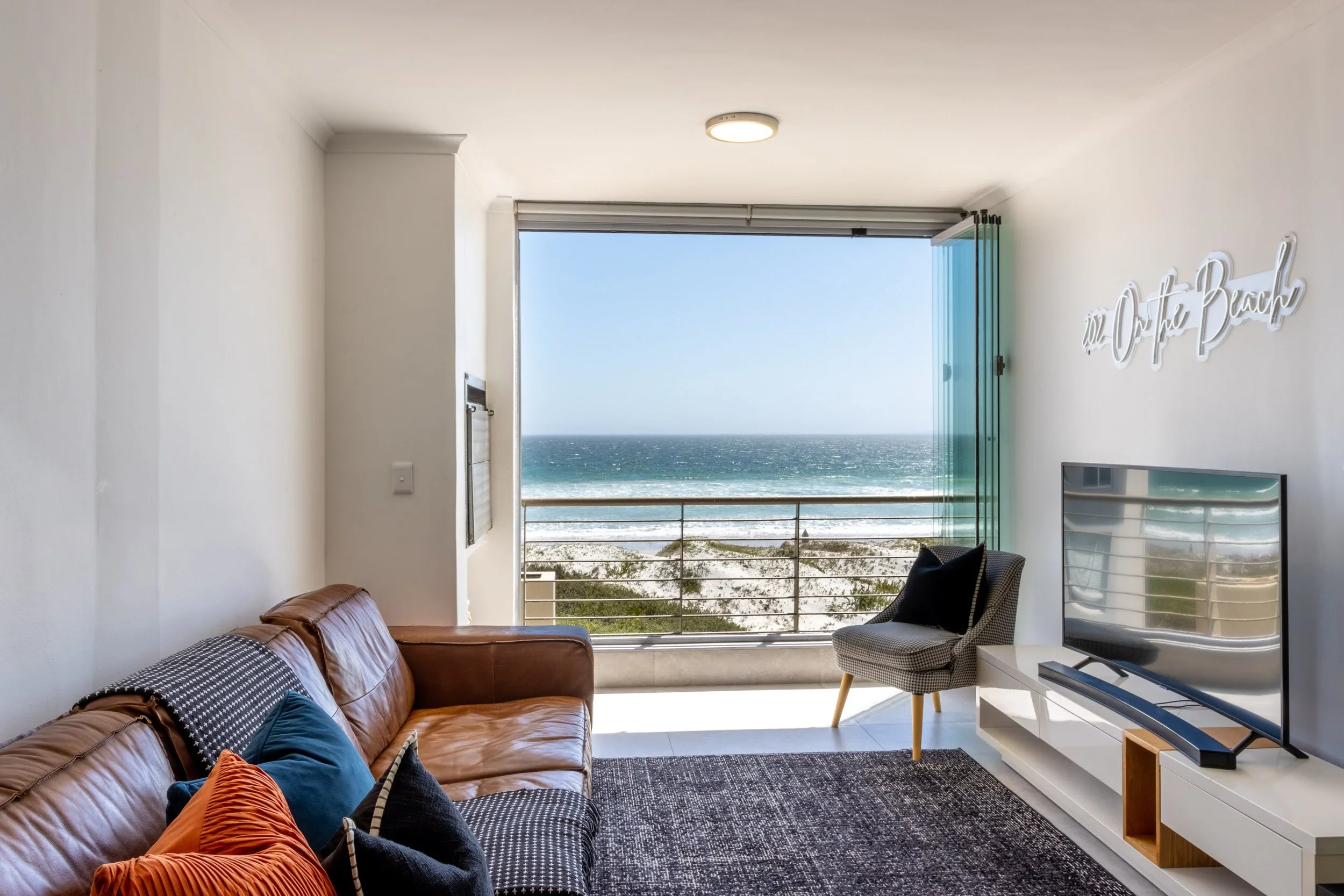 Living room with a brown leather sofa, colorful throw pillows, a patterned dark rug, a TV on a white stand, and large windows opening to a balcony with a view of the beach and ocean.