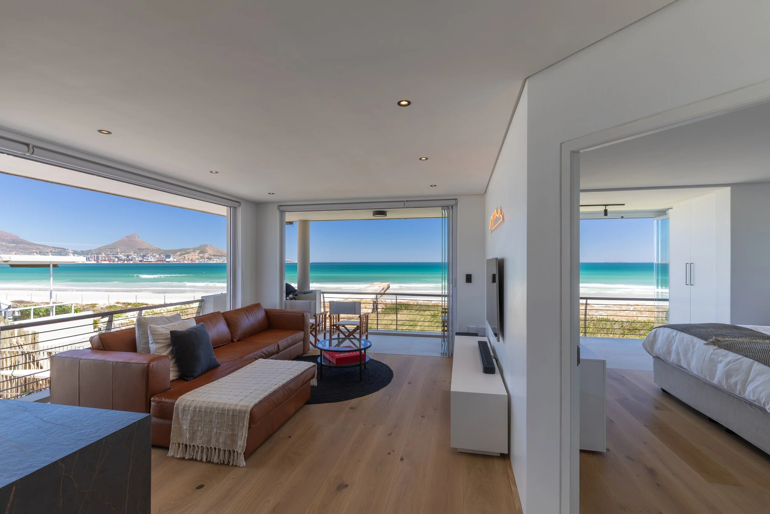 Modern living room with large windows showing a beach and mountains, tan leather sofa with pillows, small round black rug, and a view of the bedroom with a bed and ocean in the background -  Cape Town, South Africa