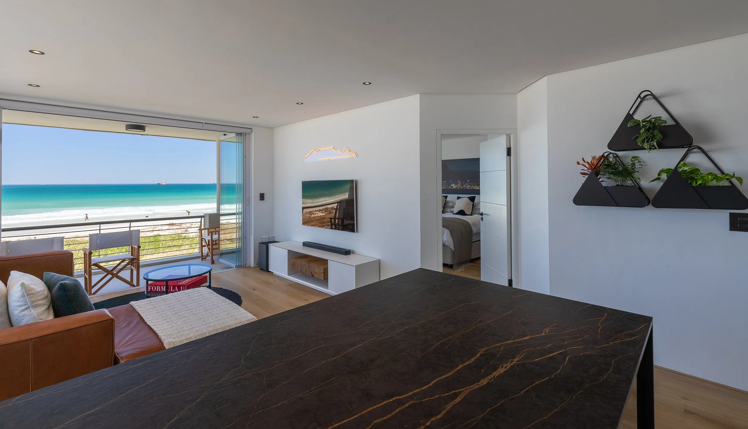 Living room on the beach with sea view through large sliding glass doors, a TV on a white stand, wall-mounted plant shelves, a bed visible through an open door, and seating furniture near the window. - Cape Town, South Africa