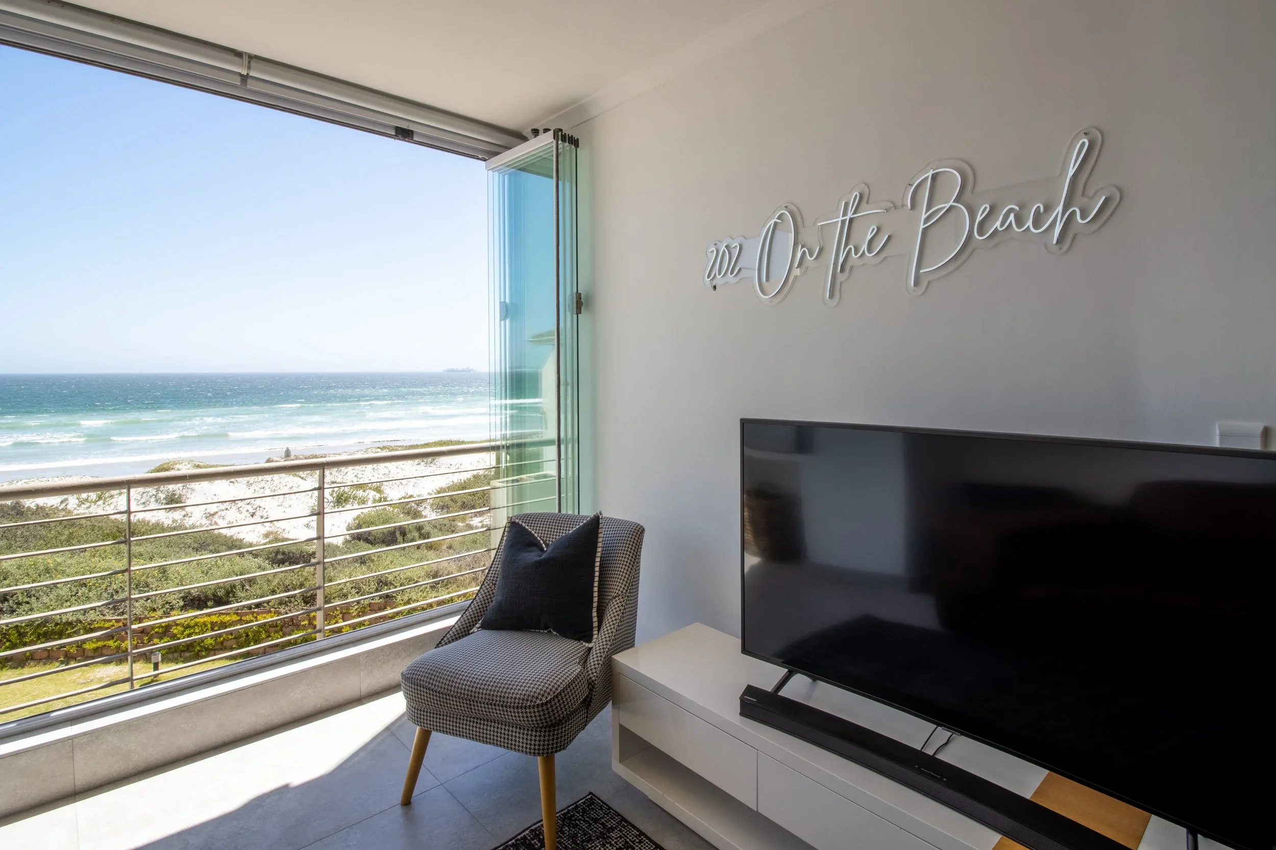 Living room with large window overlooking the beach, a chair next to a TV and a neon sign on the wall that reads 'oz On the Beach'.